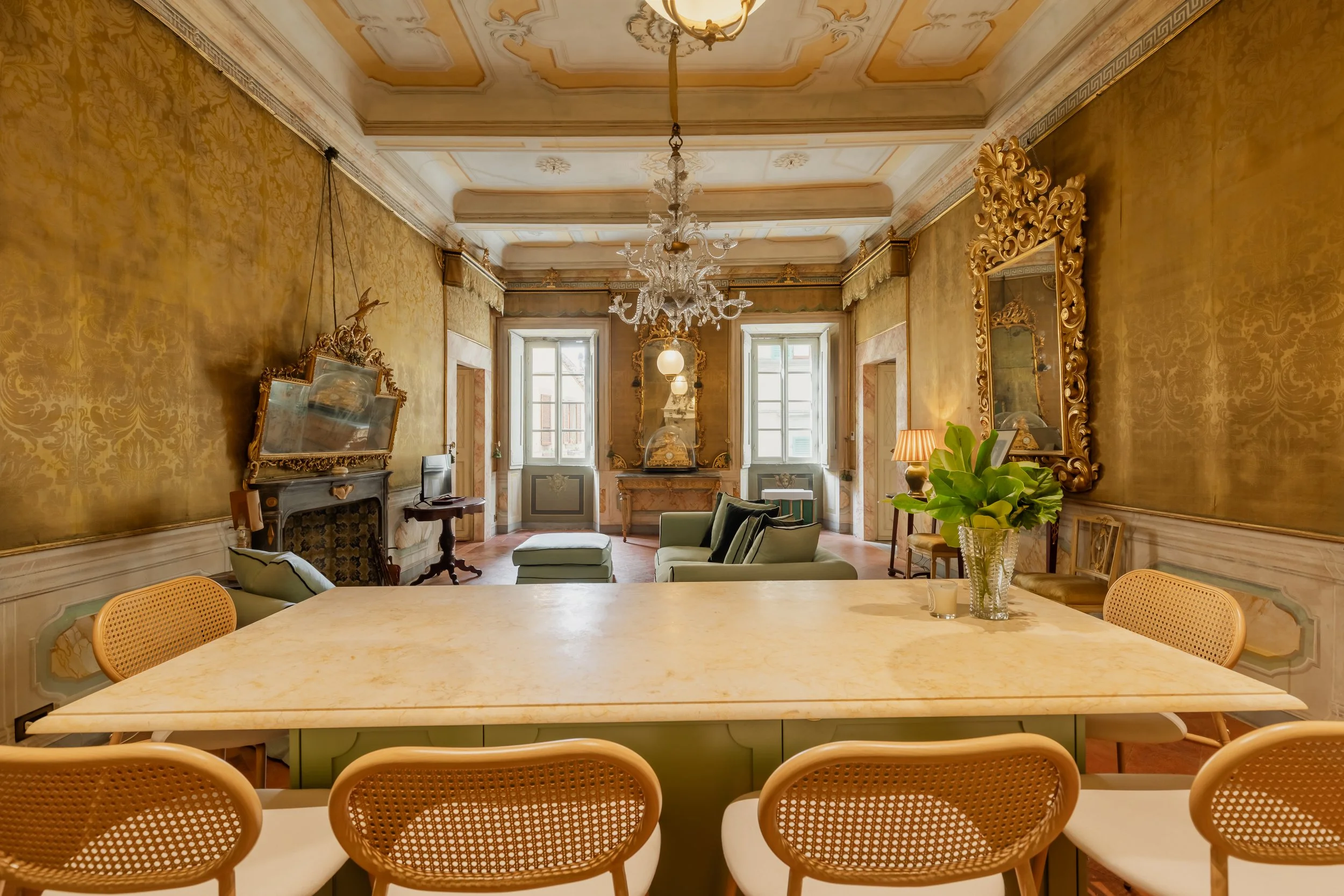 Elegant living room with a marble dining table in the foreground, surrounded by light-colored chairs. The room features ornate gold wallpaper, large windows, and classical decor including mirrors and a chandelier.