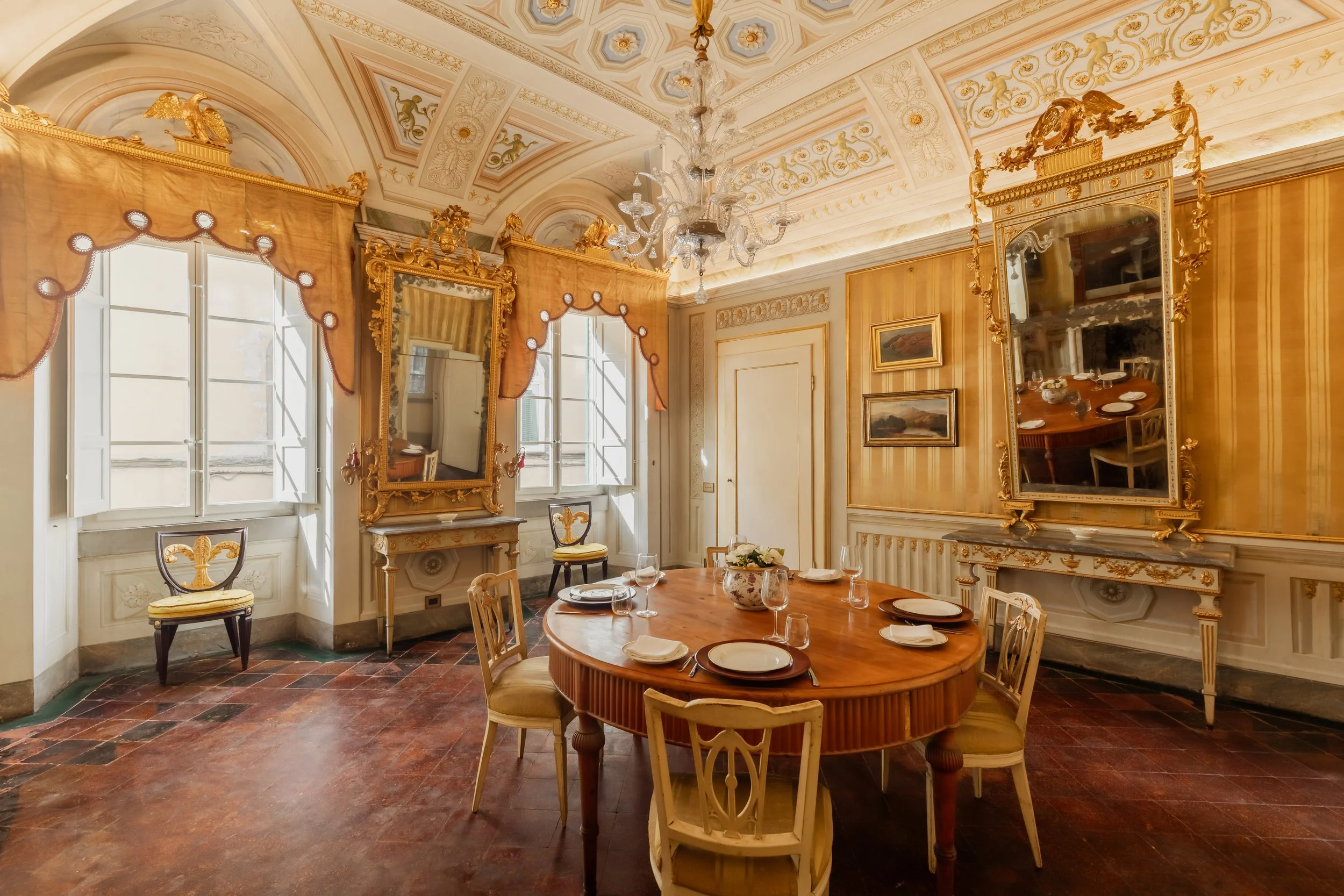 Elegant dining room with a round wooden table set with plates, glasses, and a flower arrangement, surrounded by chairs, ornate gold-framed mirrors, paintings, chandeliers, and tall windows with curtains, in a luxurious classical style.