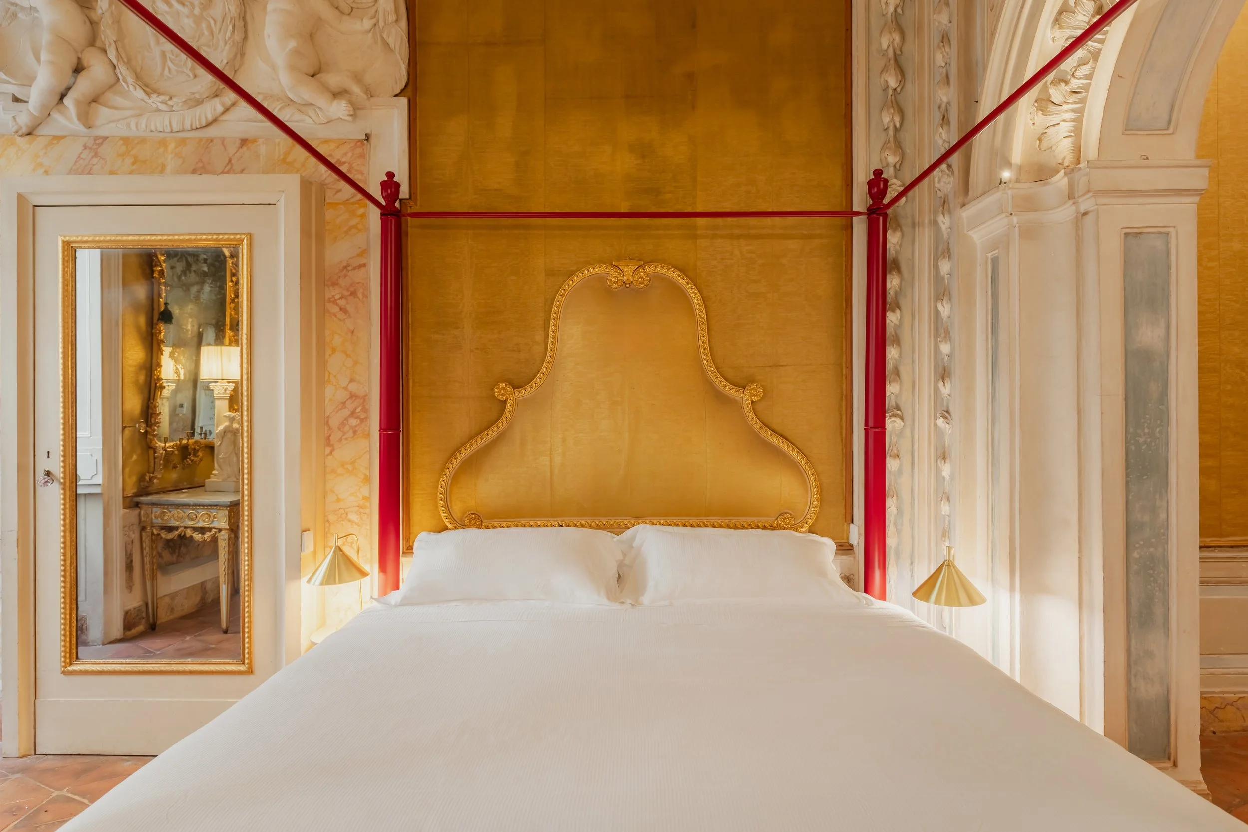 A luxurious hotel room with a white bed, ornate gold wood headboard, and red curtain rods. A full-length mirror on the left reflects a decorated vanity table with a mirror and lamp, and a wall sculpture with detailed carvings is visible above the mir