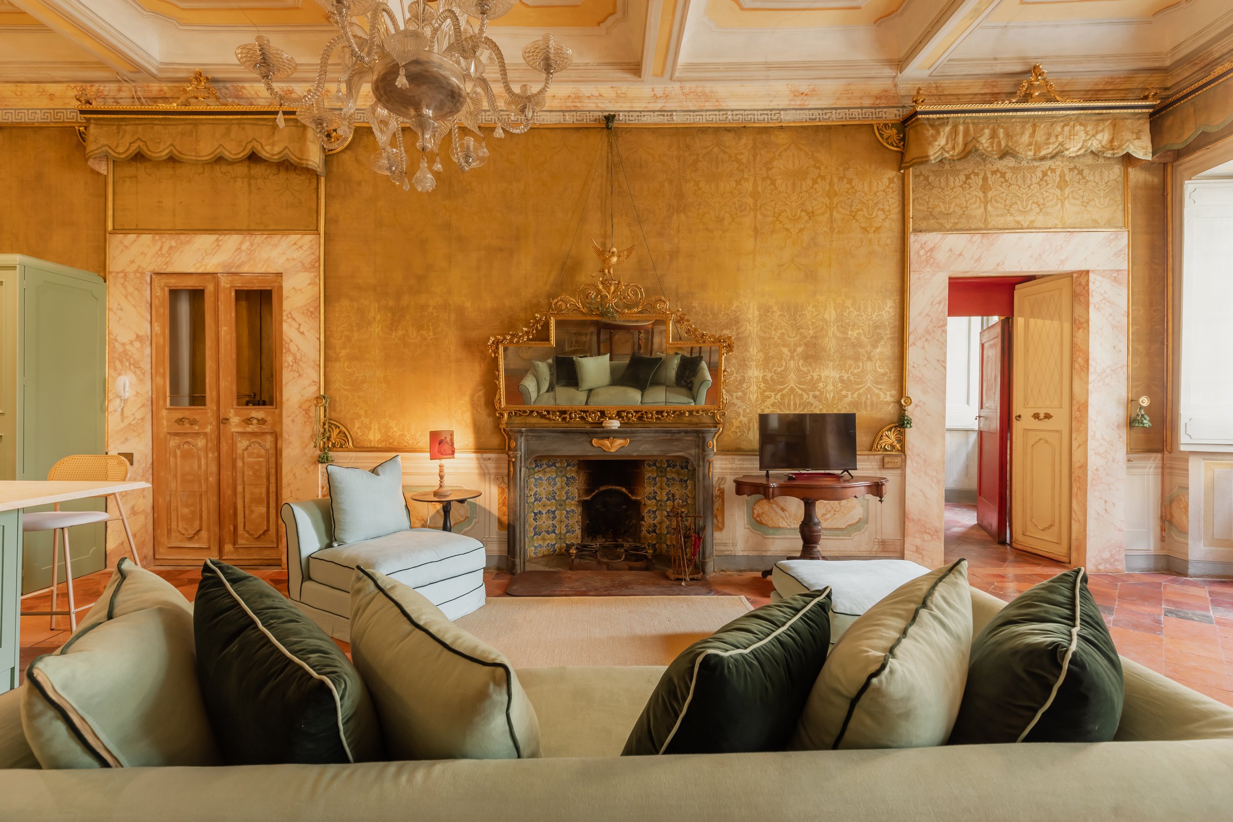 A vintage living room with ornate gold wall coverings, a marble fireplace with a mirror above, a chandelier, sofas, a small TV, and decorative pillows.