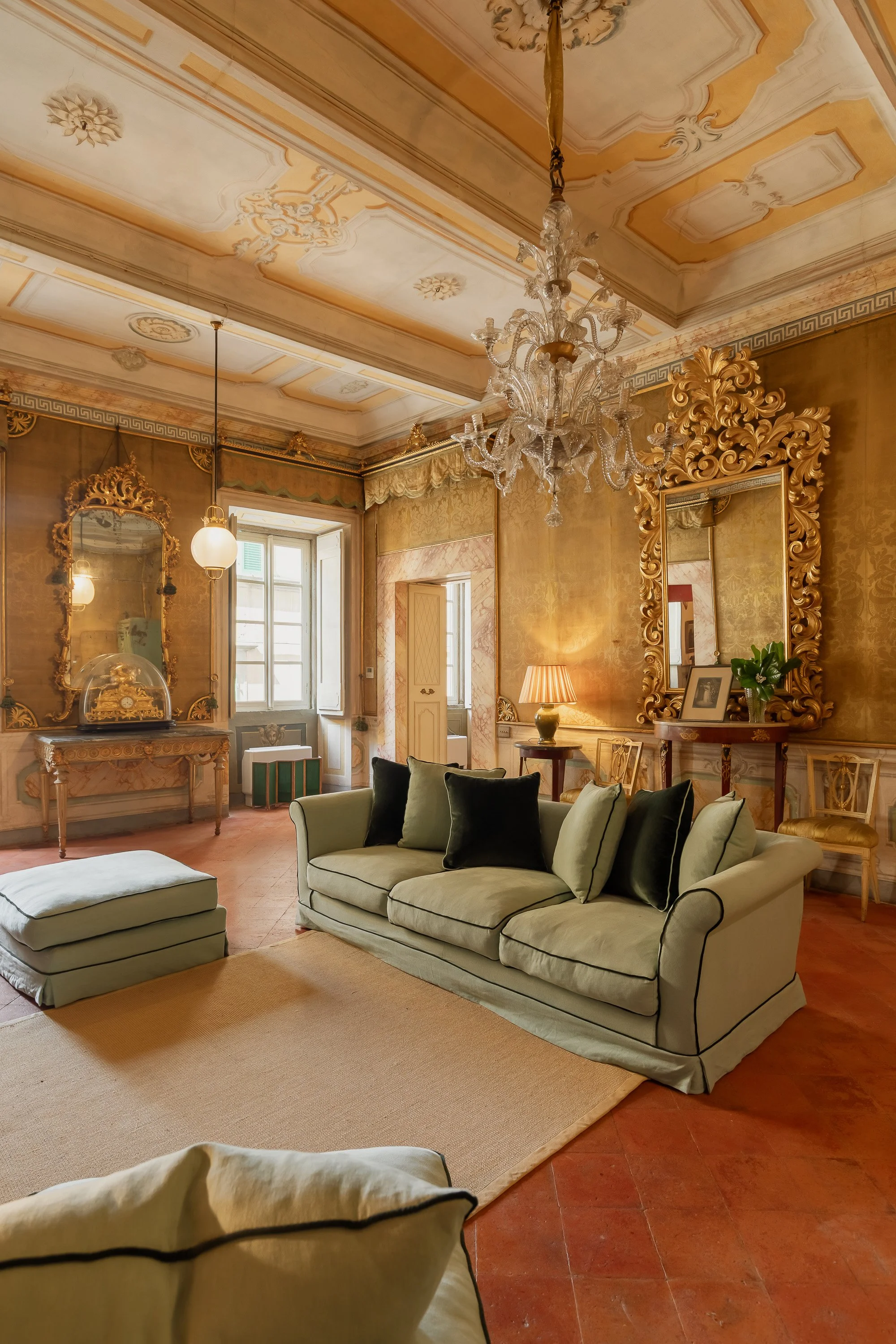 Elegant living room with ornate gold accents, a chandelier, green sofa with black and green pillows, a large mirror with a gold frame, and a window letting in natural light.