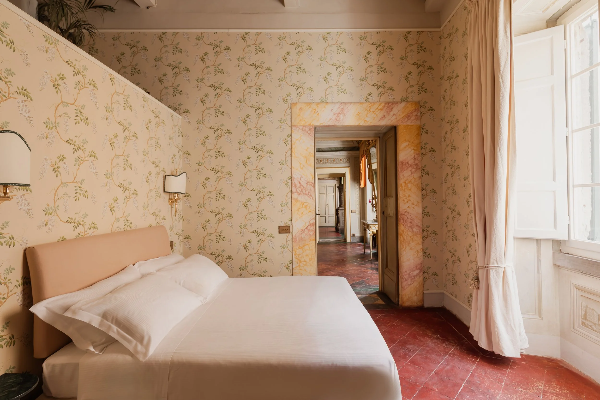 A bedroom with floral wallpaper, a beige bed with white pillows, a window with white curtains, and a doorway with a marble frame leading to a hallway with terra cotta tiled floor and classic decor.