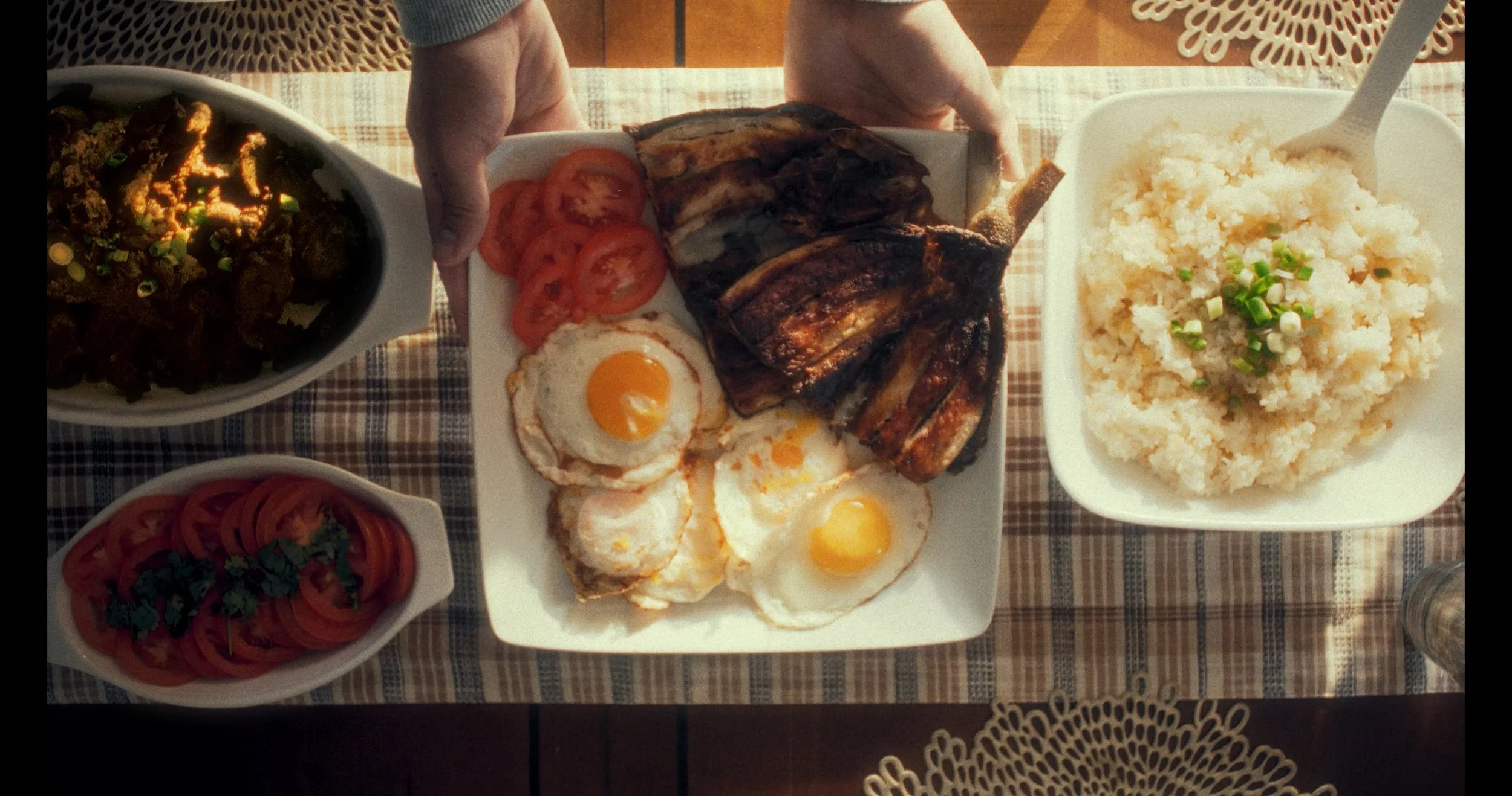 A breakfast spread featuring eggs cooked sunny side up, grilled meat, slices of tomato, mashed potatoes topped with chopped green onions, a bowl of beef stew, and a side of sliced tomatoes on a checkered tablecloth.