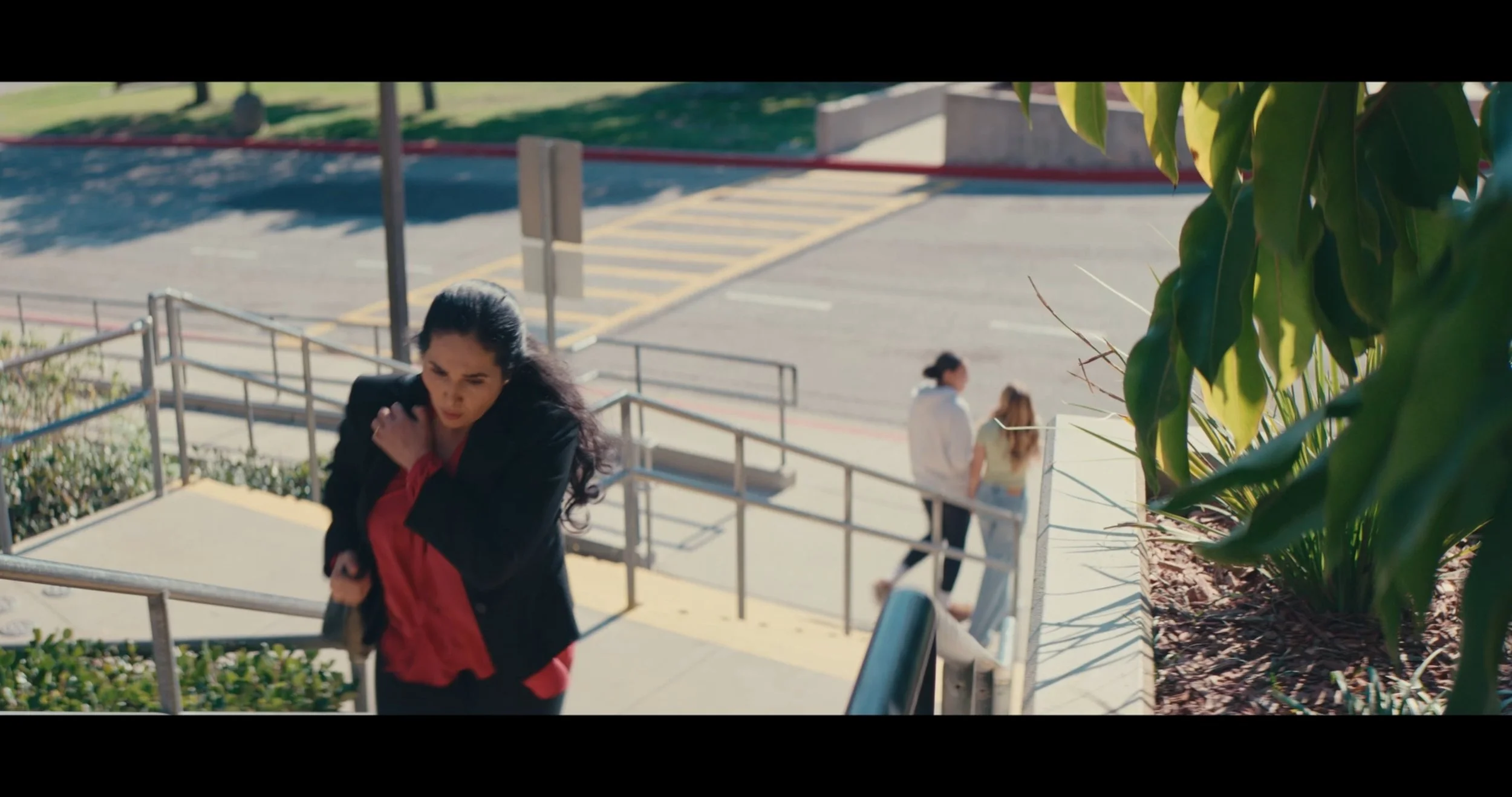 A woman with long dark hair wearing a black blazer and red shirt walking up stairs, with two women sitting on the stairs and two women walking in the background, and green plants on the right side.