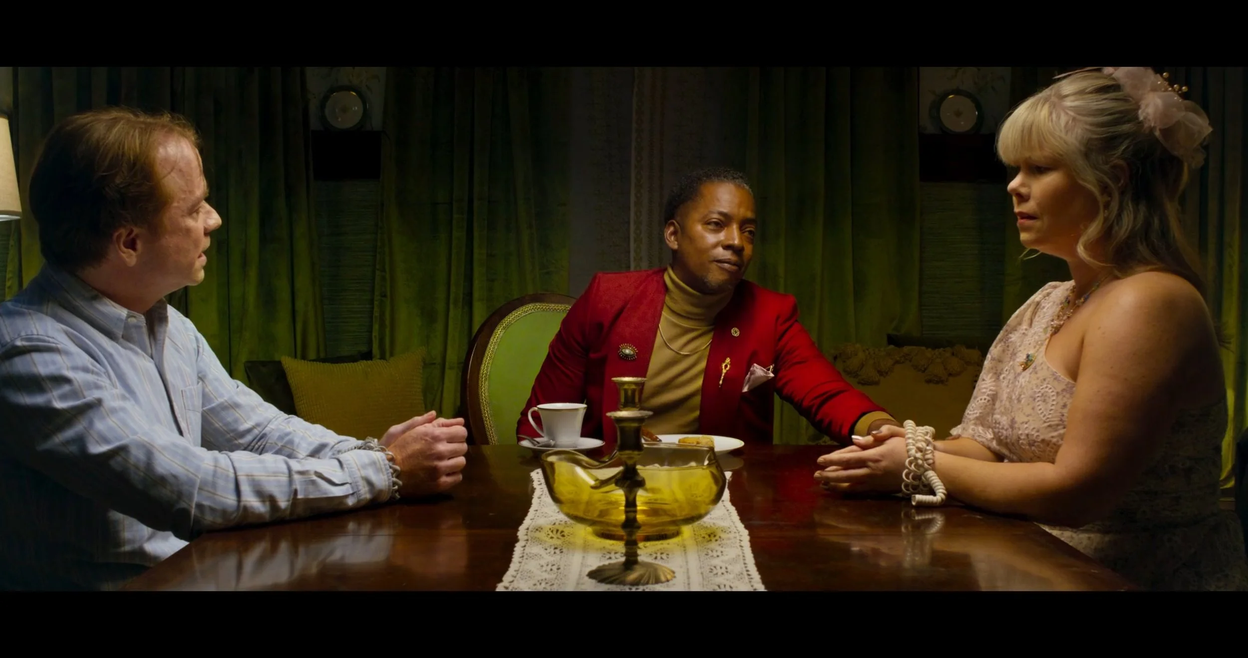 Three people sitting at a dining table, holding hands, with a man in a red jacket in the middle, a woman in a lace top on the right, and a man in a striped shirt on the left, with green curtains in the background.