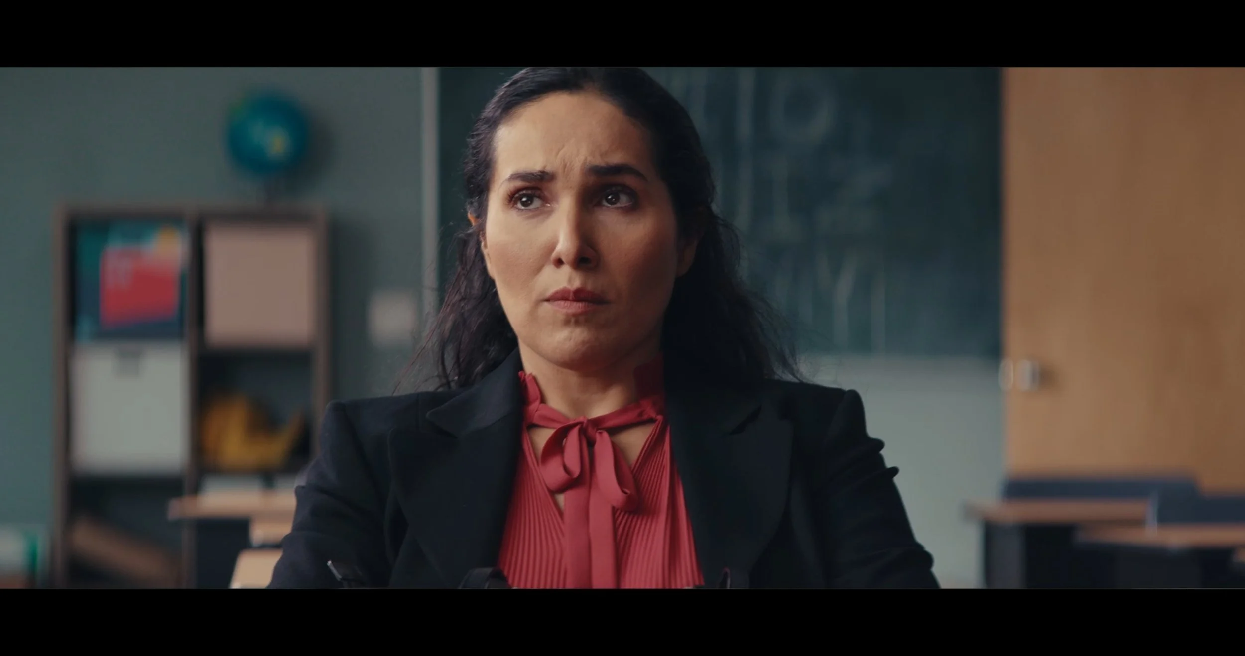Woman with dark hair wearing a black blazer and a red blouse with a bow, sitting at a desk in a classroom or office setting.
