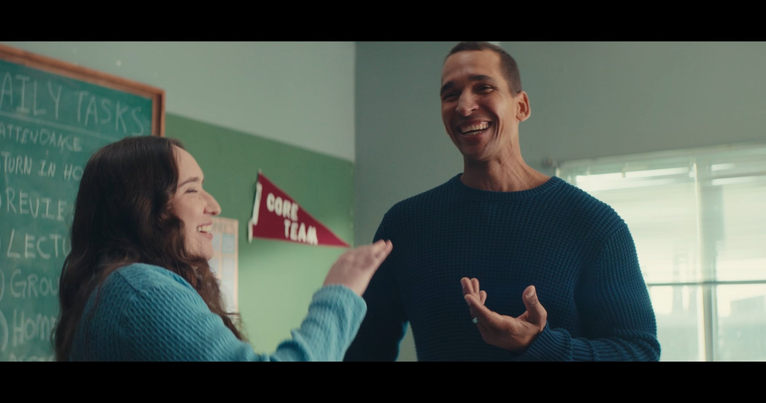 A woman and a man are smiling and talking to each other in a classroom. The woman has long dark hair and is wearing a blue sweater. The man has short dark hair and is wearing a dark blue sweater. Behind them, there is a chalkboard with writing on it 