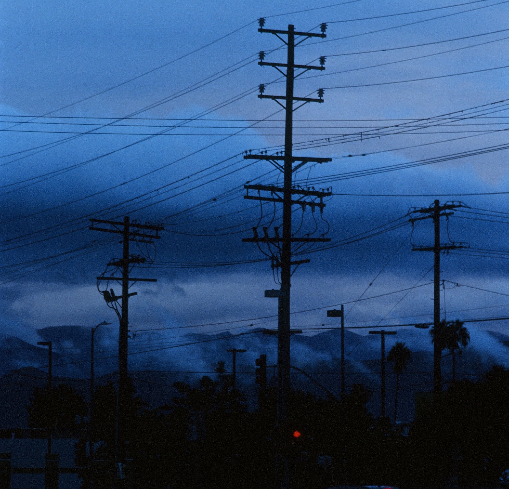 Silhouettes of multiple telephone poles and power lines against a dark evening sky with some clouds and a small red light at the bottom.
