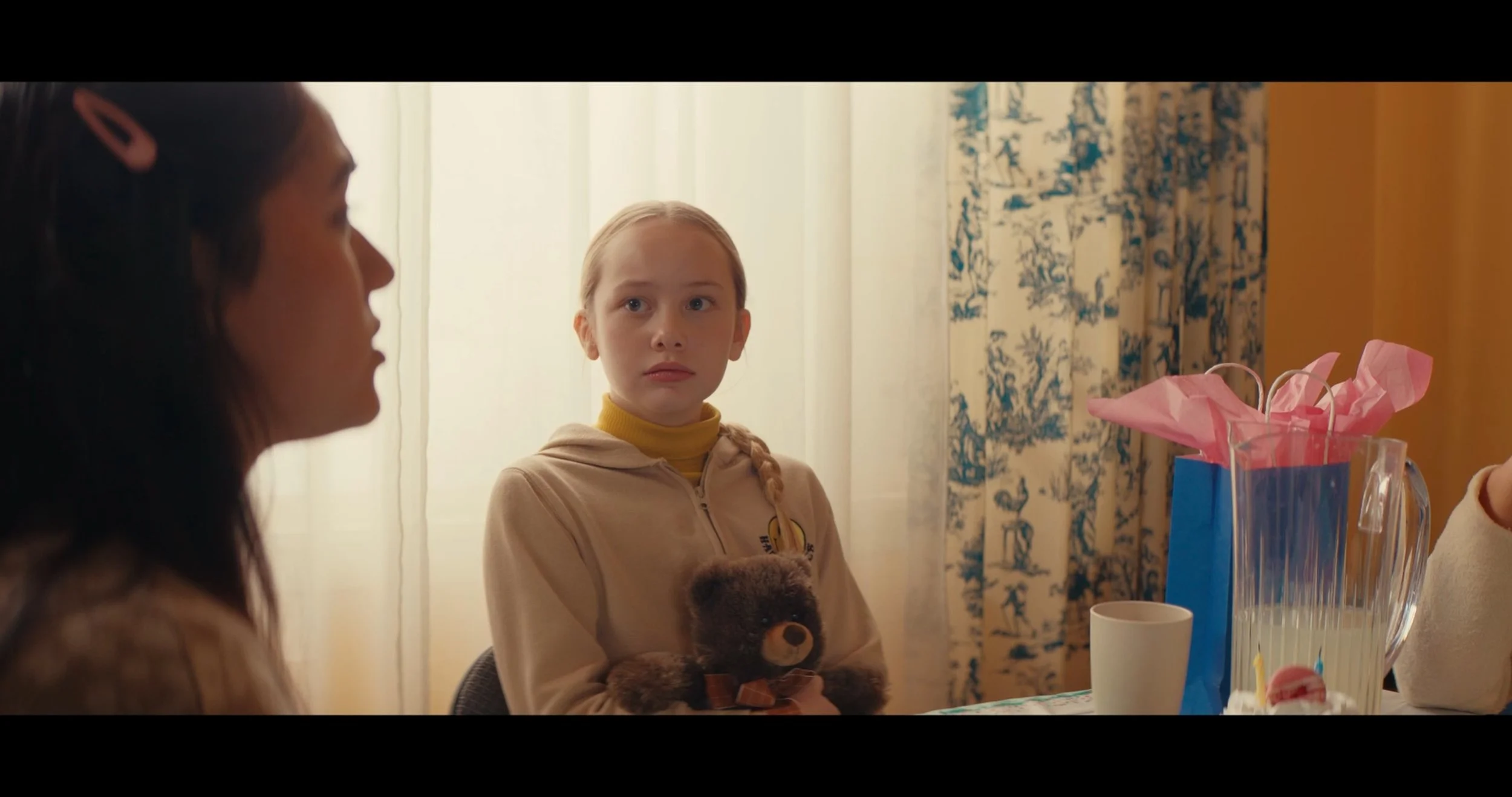 A young girl with tied back hair and a beige hoodie, holding a teddy bear, looking at another woman in a room with floral curtains and a yellow wall, with gift bags and a poured drink on the table.