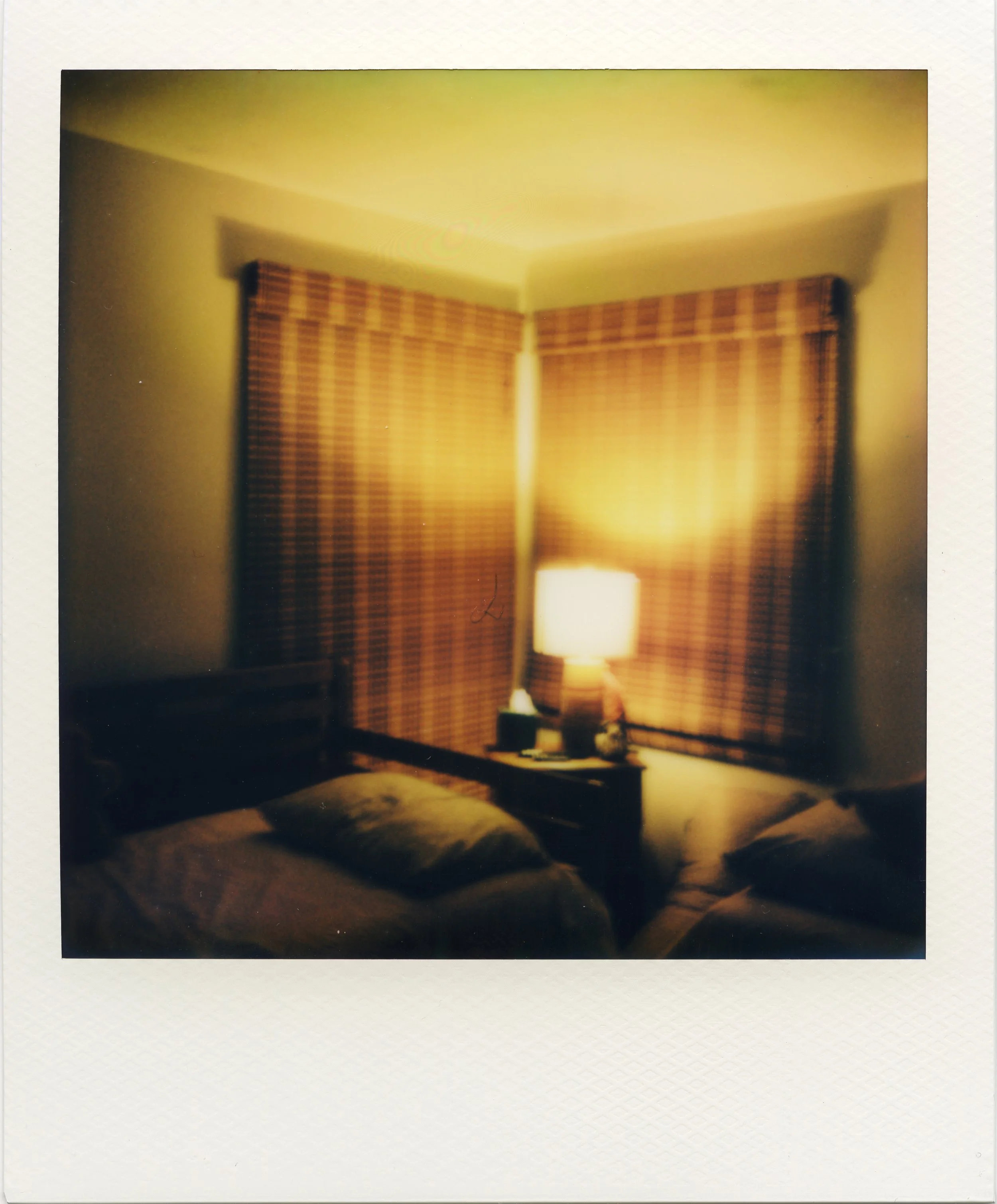 A cozy bedroom with a lamp on a nightstand, covered with a blanket, and windows with closed wooden blinds at night.