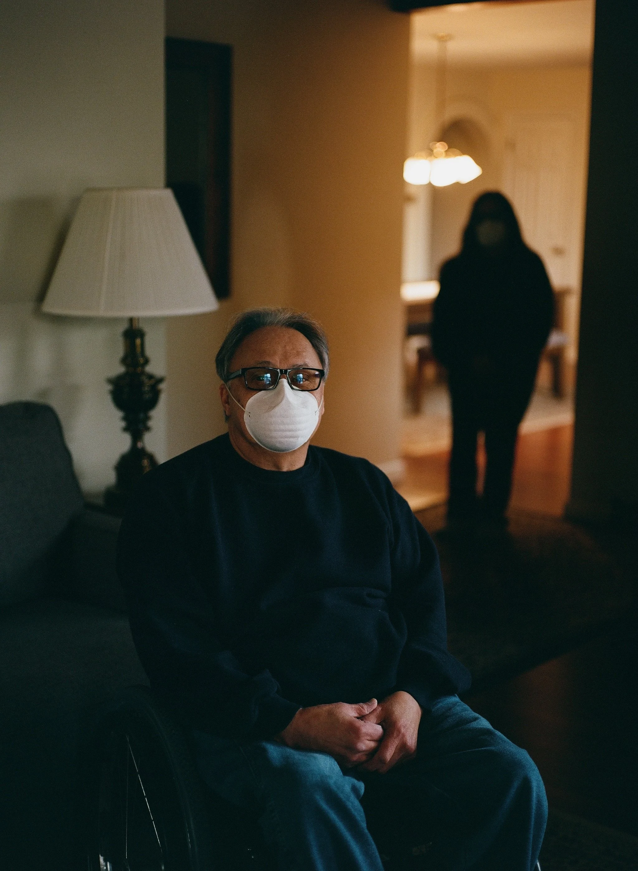 A man with glasses and a white face mask sitting on a black couch, in a dimly lit room, with a woman in a dark outfit and face mask standing in the background near a doorway.