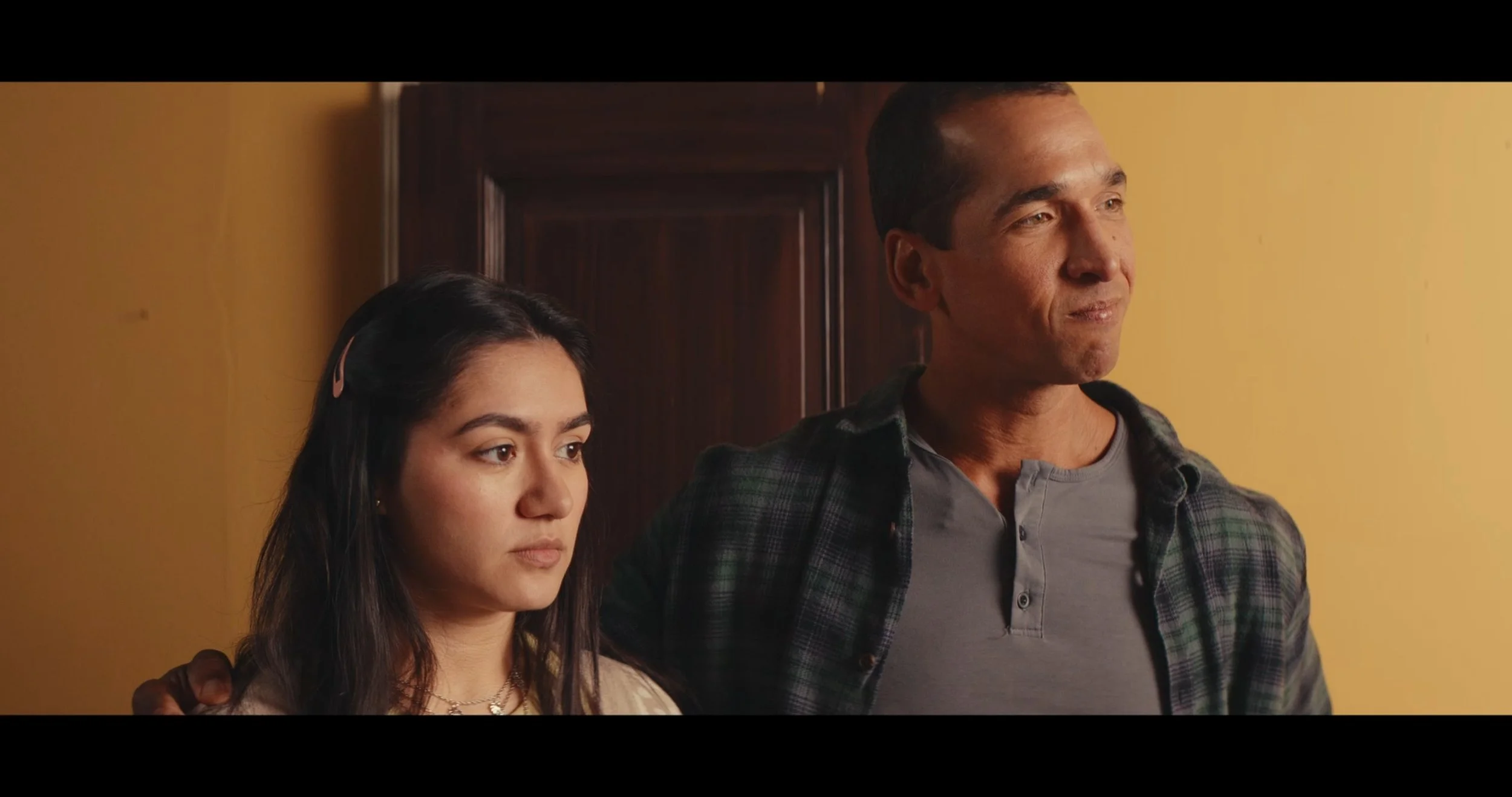 A young woman with dark hair and light skin standing next to a man with short hair and a plaid shirt, both looking to the right with neutral expressions, indoors with yellow walls.