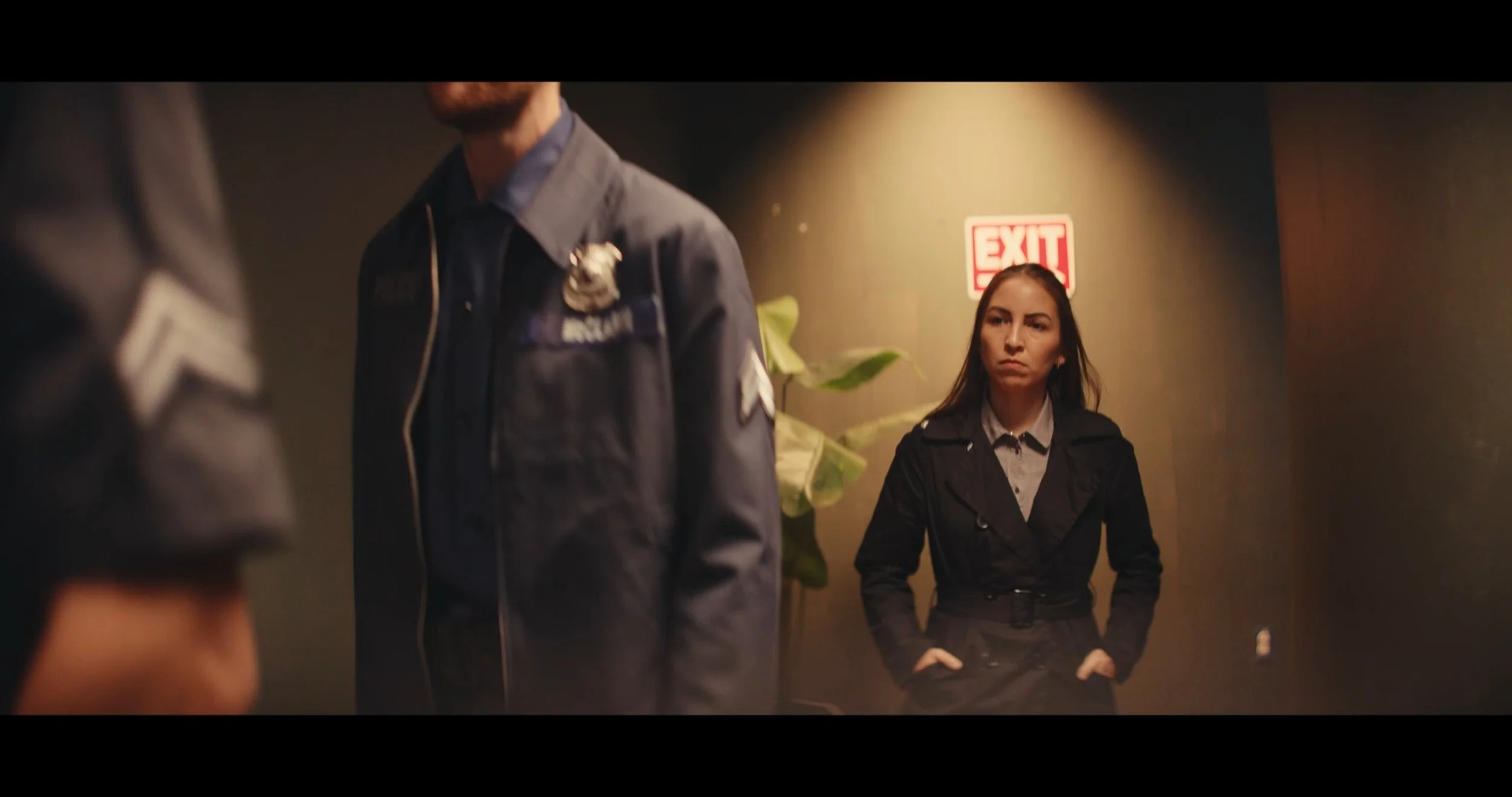A woman with long hair, wearing a black jacket and gray shirt, standing with her hands on her hips behind a person in front of her. The background shows a beige wall and a red exit sign.