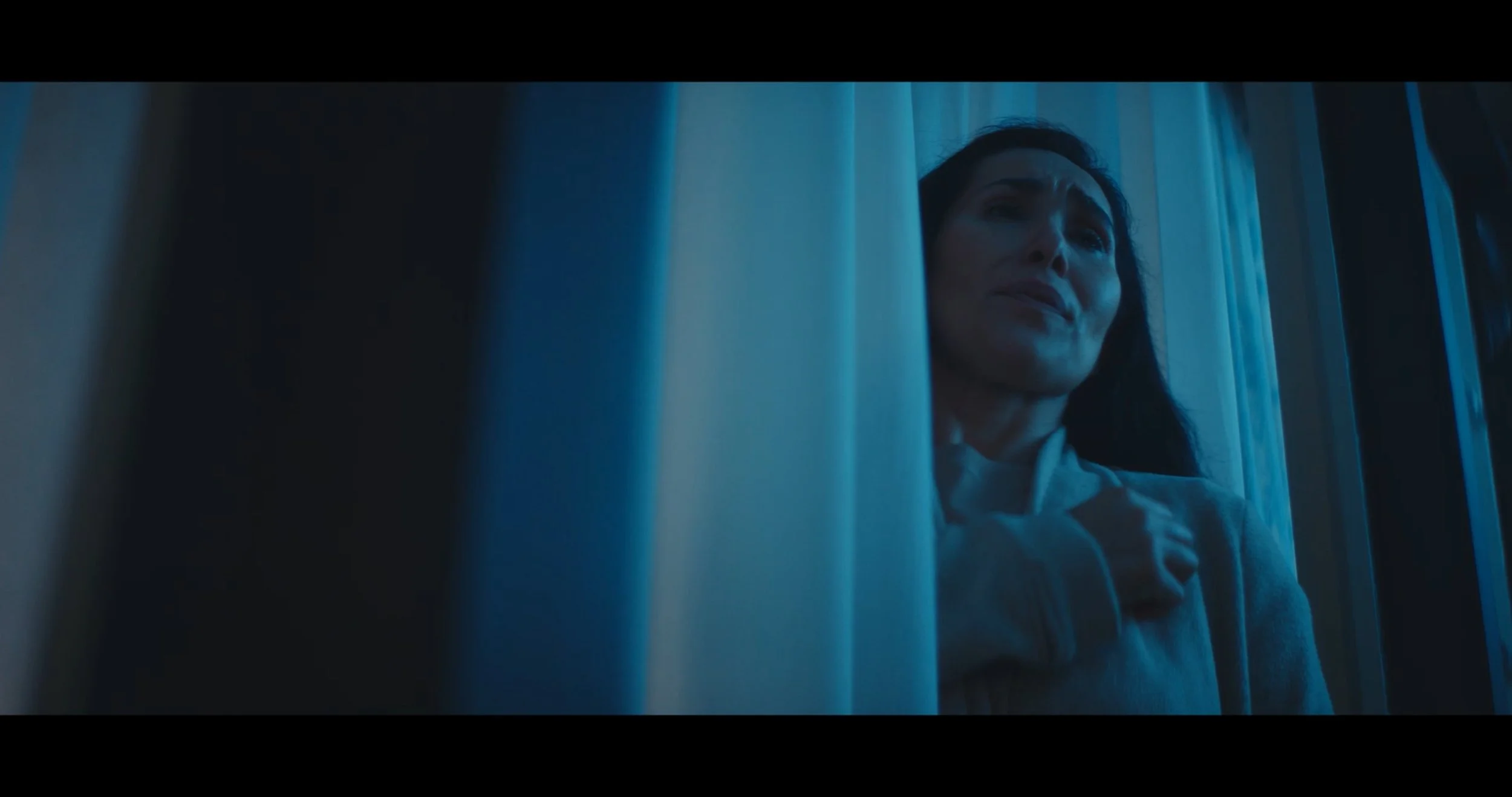 A woman with dark hair looking out a window with a somber expression, illuminated by blue light.