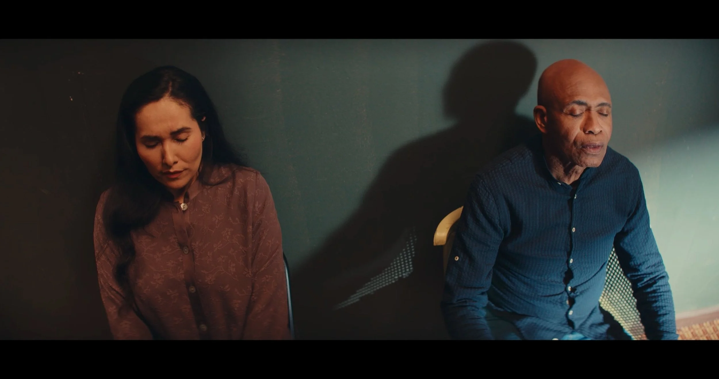 A woman and a man sitting with eyes closed, seemingly meditating or resting against a teal wall, with shadows cast on the wall behind them.