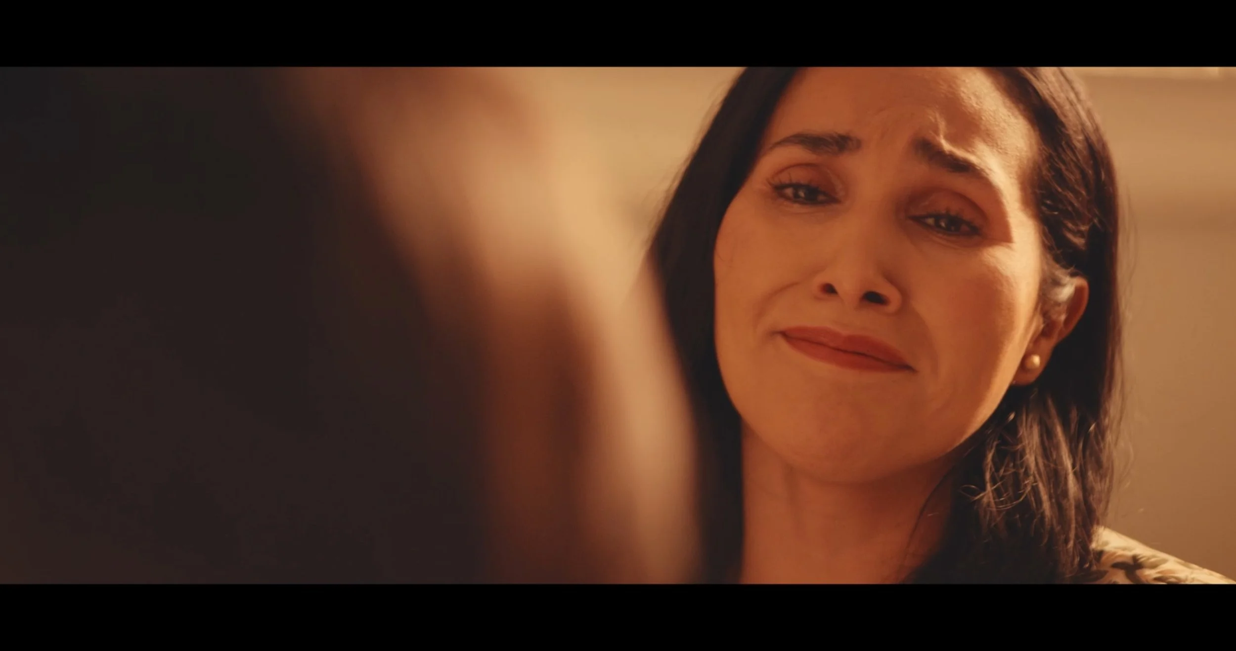 A woman with long dark hair and earrings is looking at someone out of focus in front of her, smiling softly.