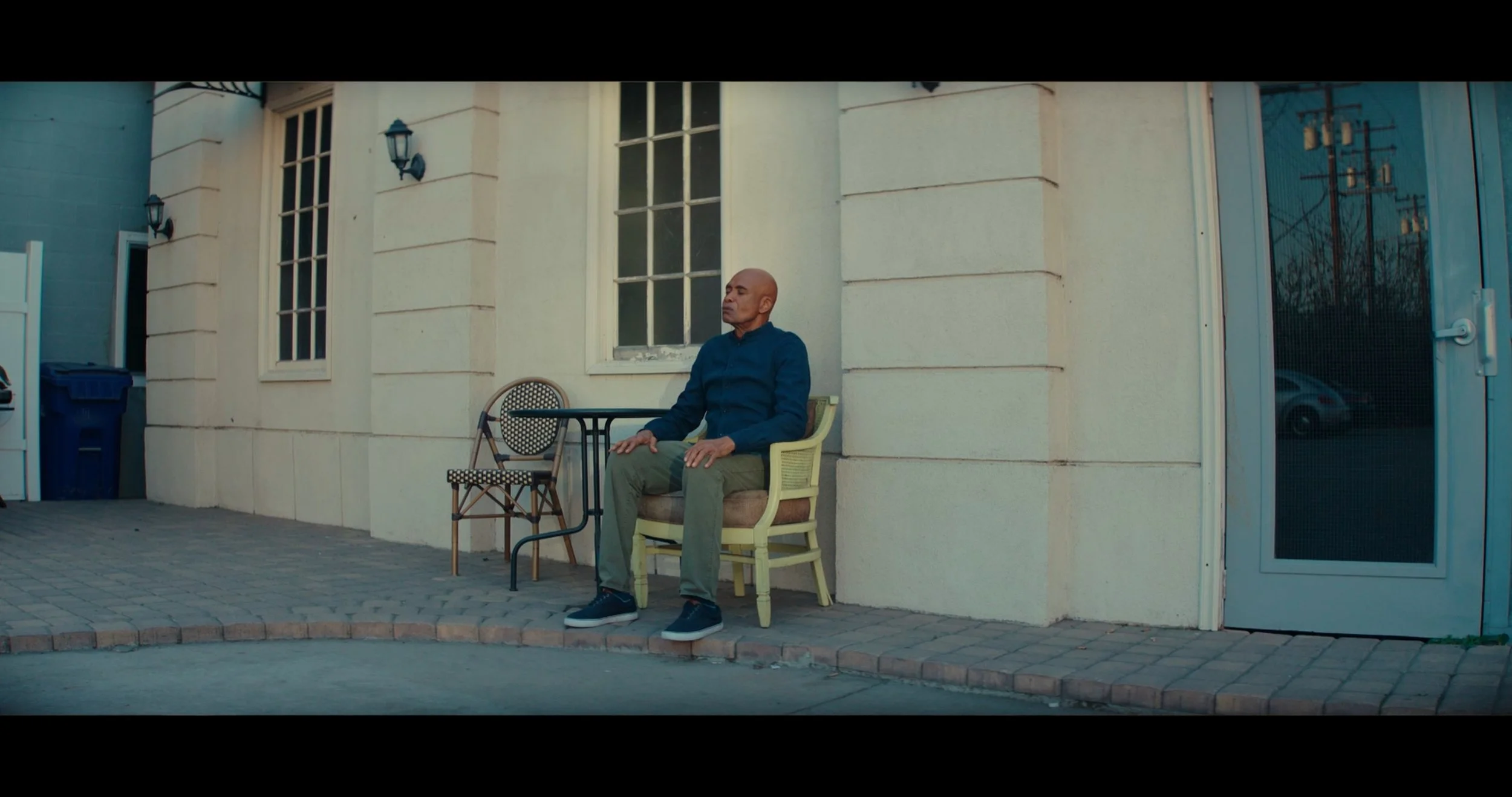 A man sitting alone on a yellow chair outside a building with large windows and beige walls, next to a small metal table and a patterned chair.