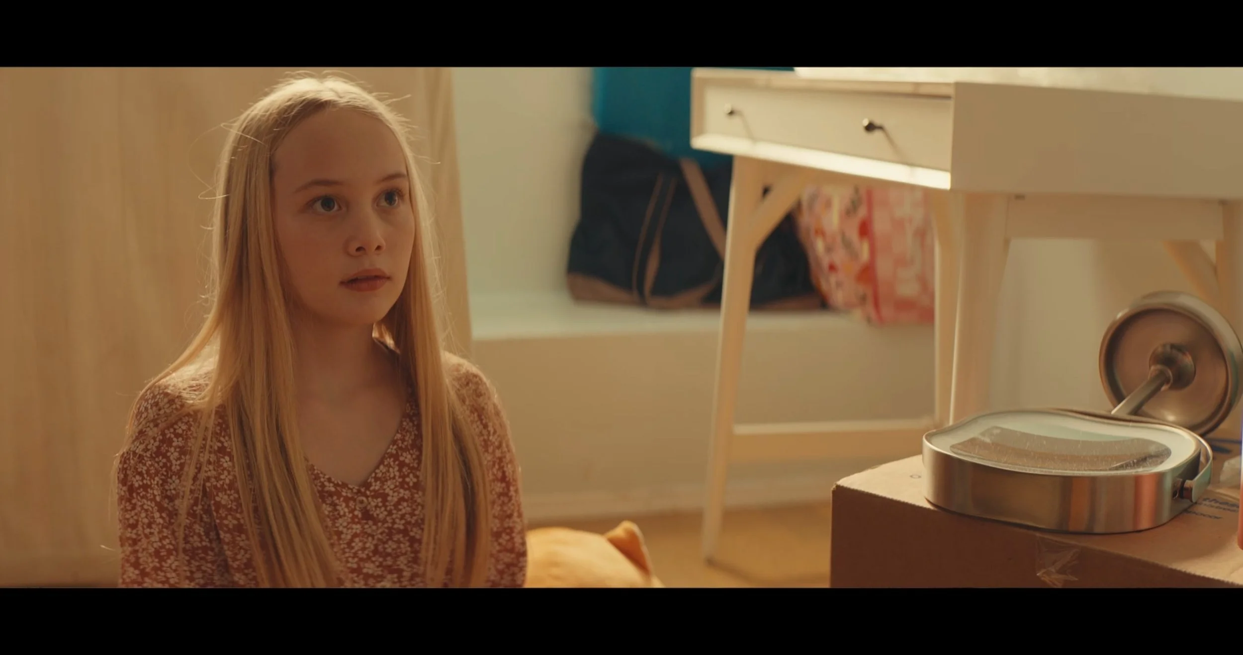 A young girl with long blonde hair sitting in a room, looking at a robotic device on a table.