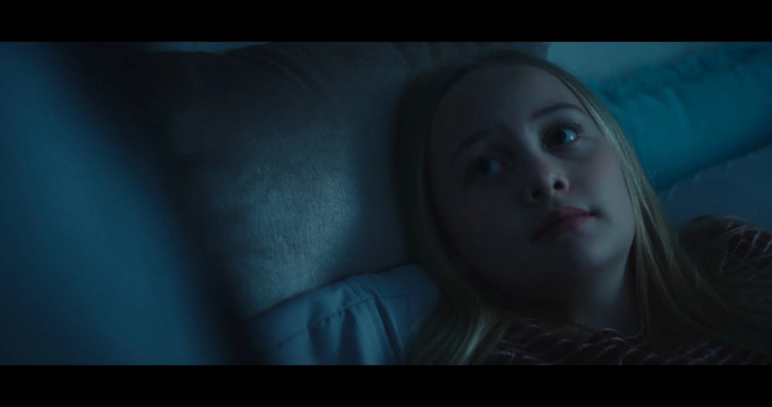 A young girl with long blonde hair and a serious expression lying on a bed in a dimly lit room.