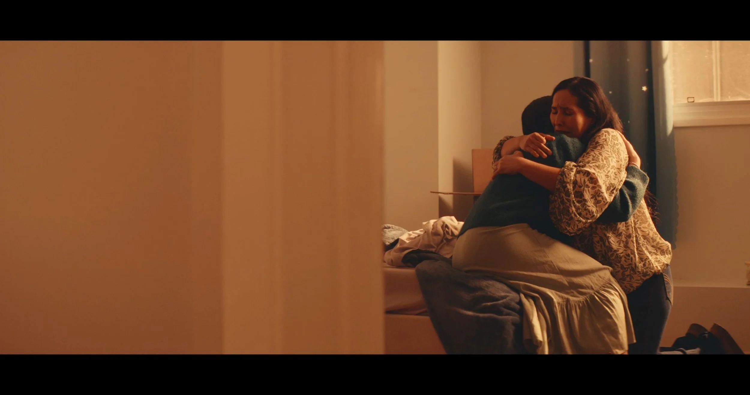 A woman and a man share an embrace in a warmly lit room, with the woman visibly emotional and the man holding her closely.