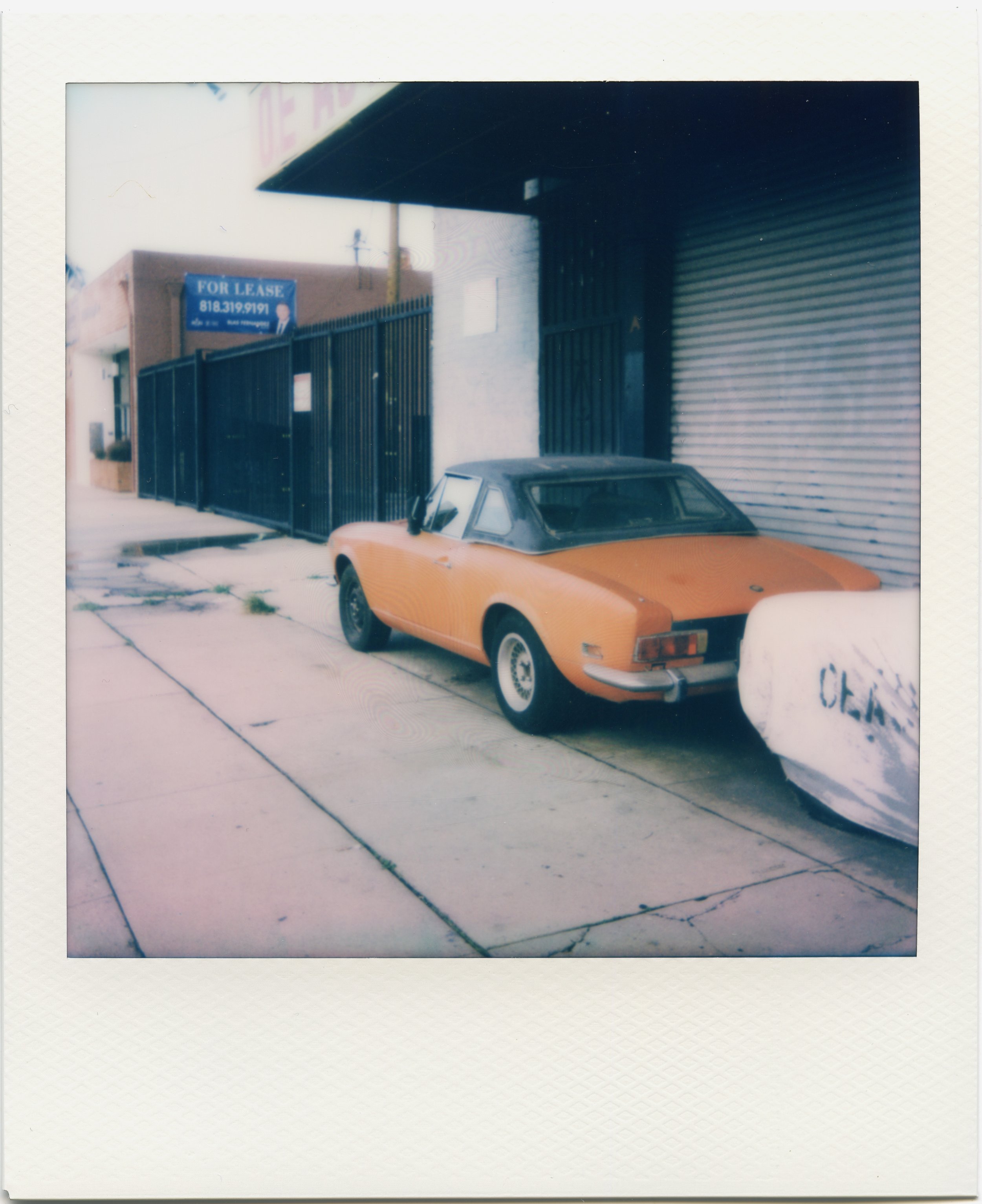 A vintage Polaroid photograph showing a yellow sports car parked on a sidewalk in front of a building with a black metal fence and a sign indicating the property is for lease. The building has a garage door, and part of the metal awning is visible.