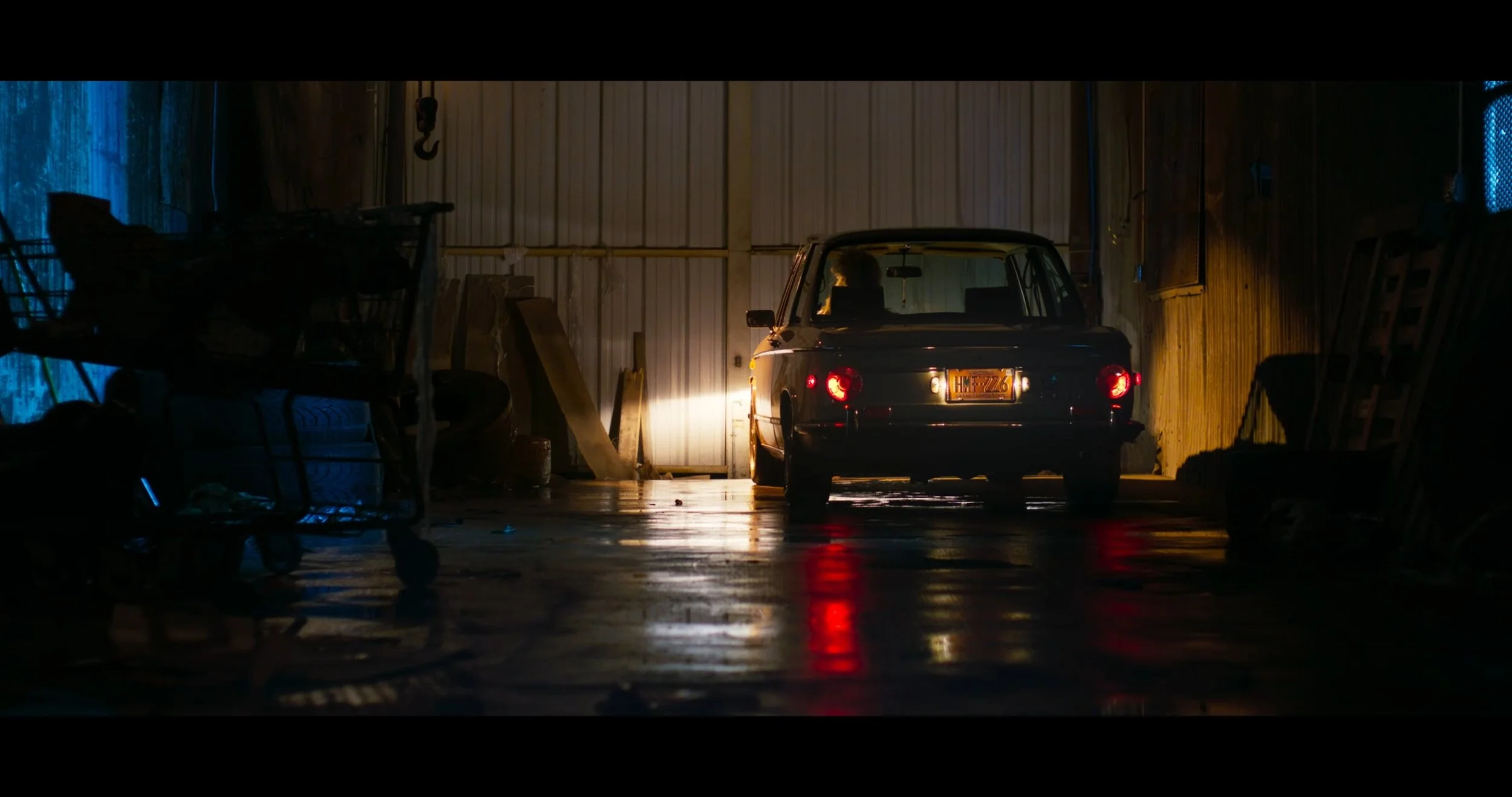 The image shows a vintage black car parked inside a dimly lit warehouse or garage. The car is illuminated by a single beam of light from the left, reflecting on the wet floor. Surrounding the car are various objects and shelves, with some blue and yellow painted walls visible.