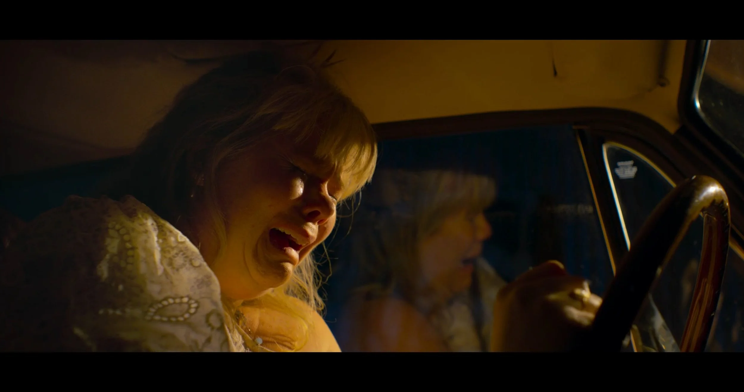A woman with blonde hair and a patterned yellow dress appears to be inside a vehicle, possibly a car, with a dark background and another person partially visible beside her.