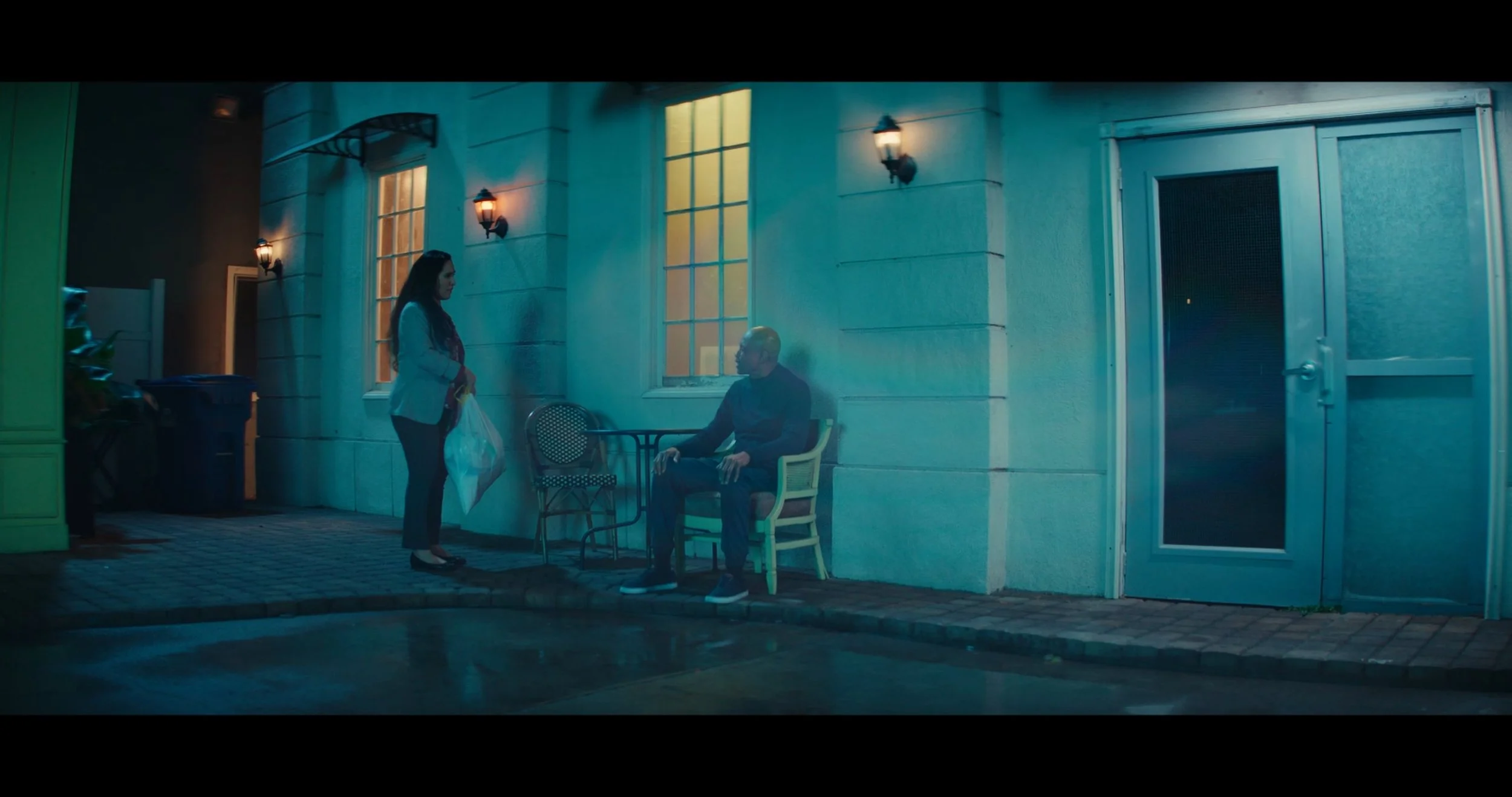 A woman is standing and handing a plastic bag to a man who is sitting on a bench outside a building at night. The building has two bright lights and large windows.