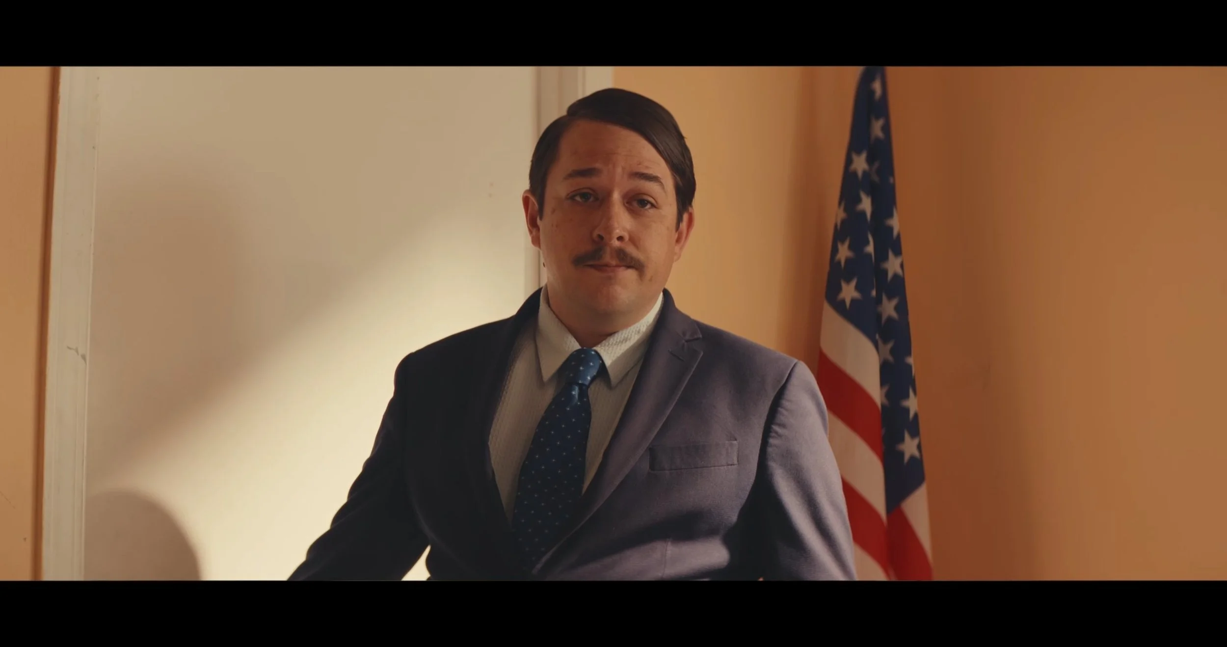 A man in a suit with a mustache, standing in front of an American flag inside a room with beige walls.