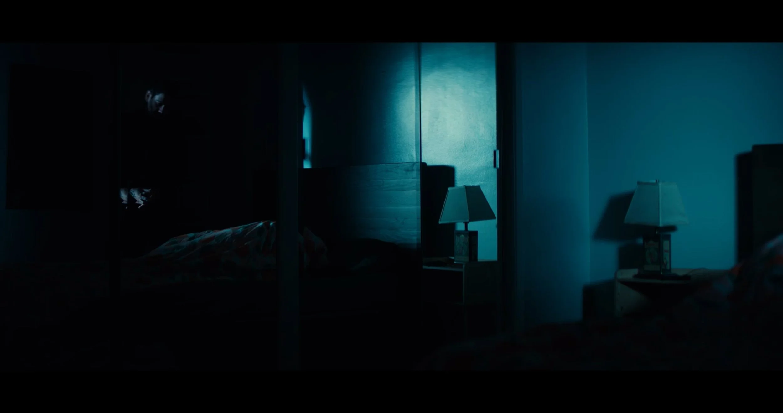 Darkly lit bedroom with two bedside tables and lamps, bed partially visible on the left, and a person standing in the shadows on the far left.