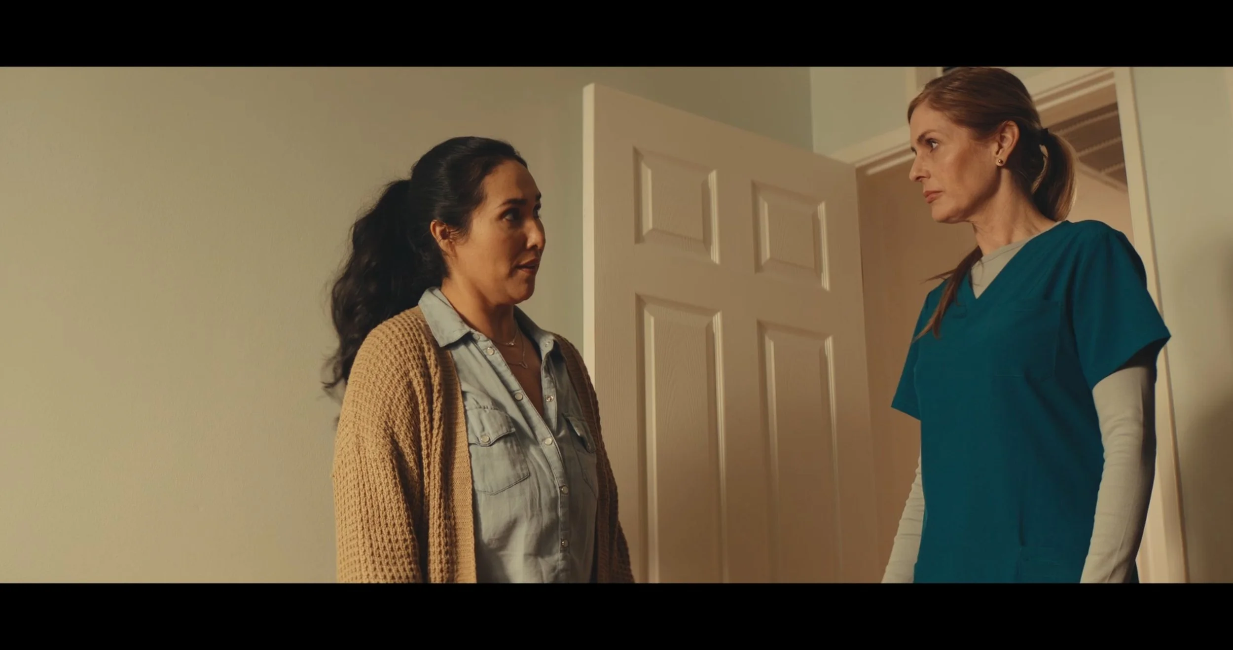 Two women standing face to face near a partially open door indoors, engaged in conversation. One woman is wearing green scrubs, and the other woman is wearing a casual jacket over a shirt.
