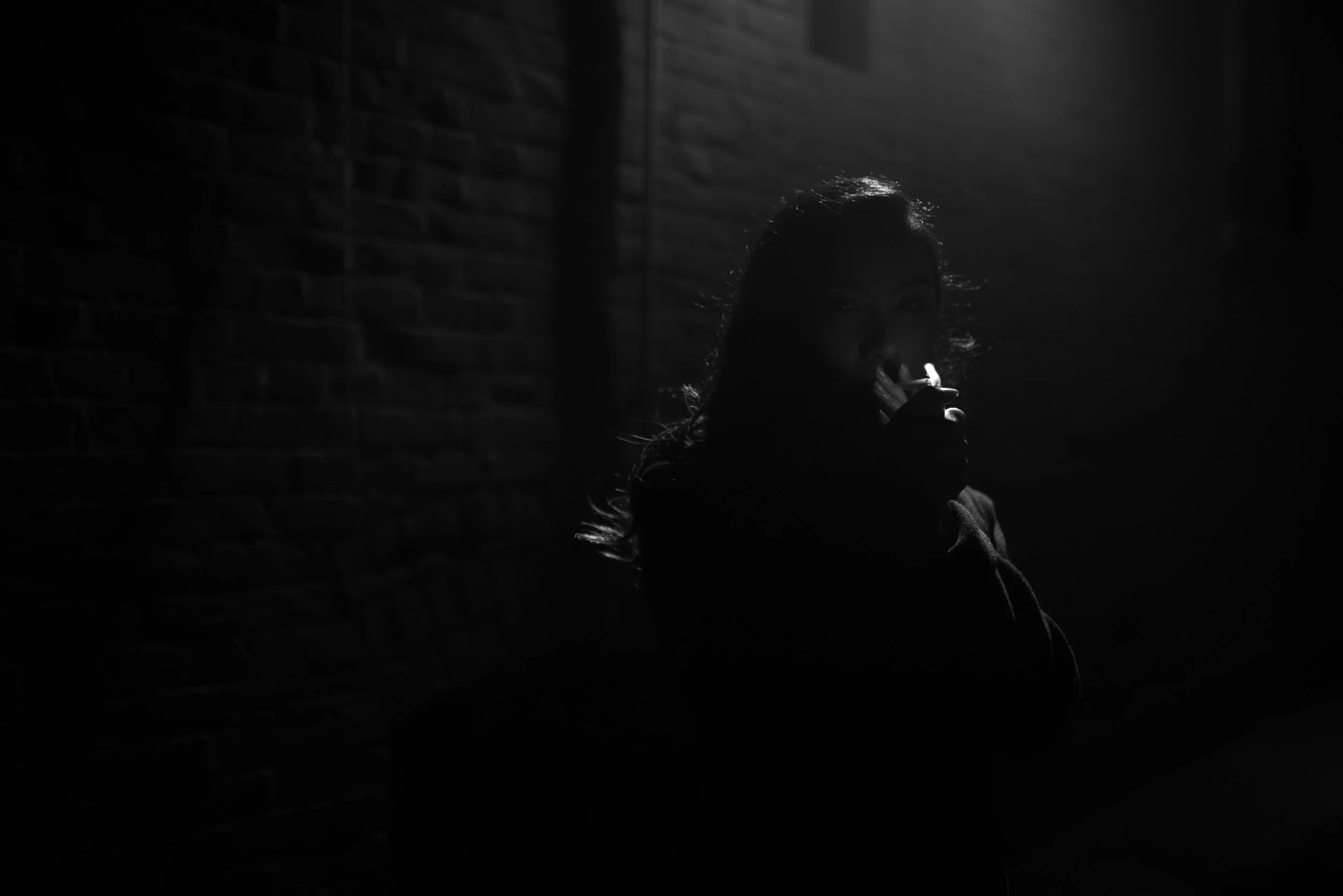 A person with long hair is partially visible in a dark, shadowed environment, holding a cigarette near their mouth. The background features a brick wall. The lighting is low and dramatic.