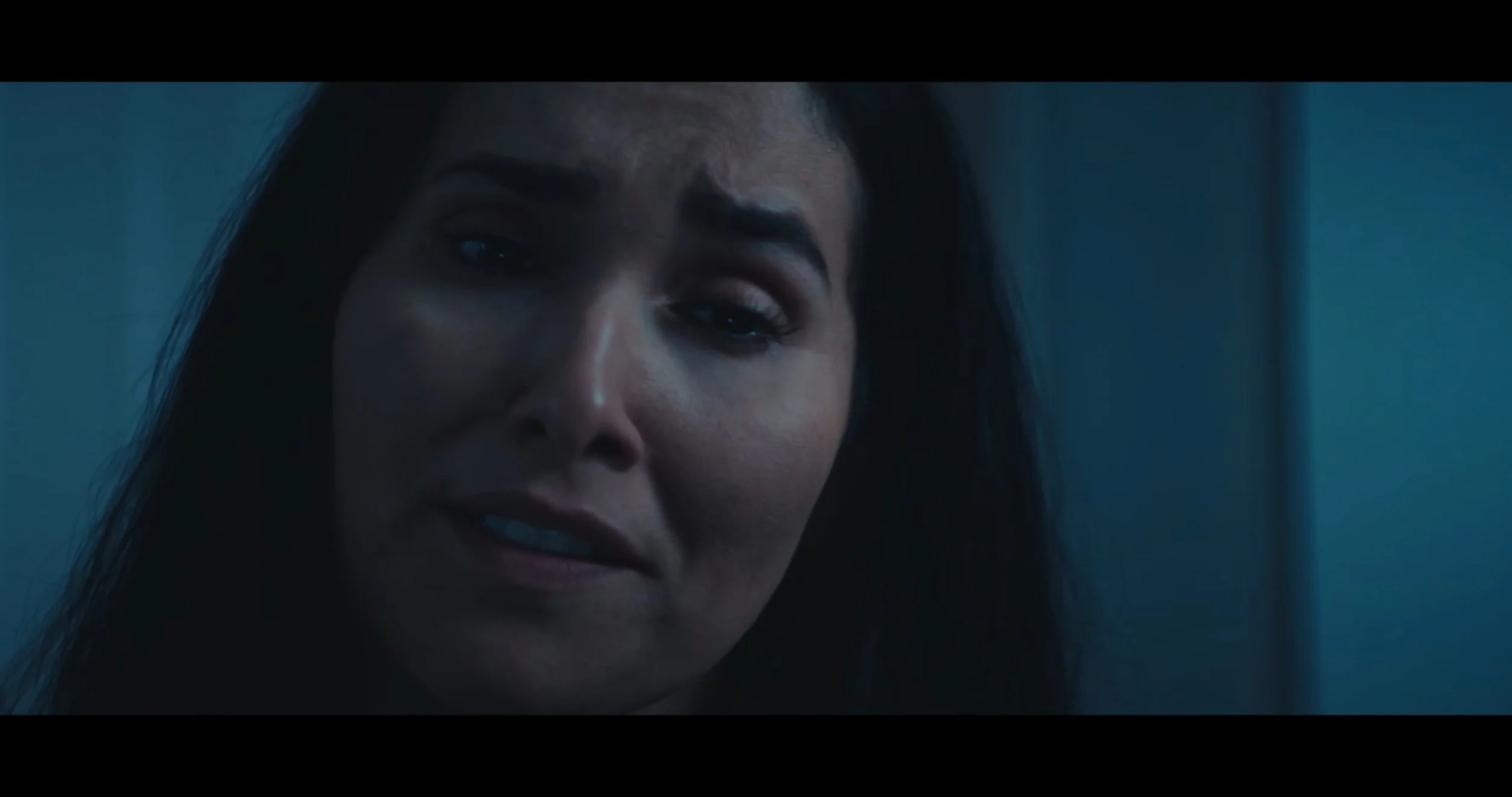 Close-up of Magdalena, smiling softly, in a dimly lit room with blue tones.