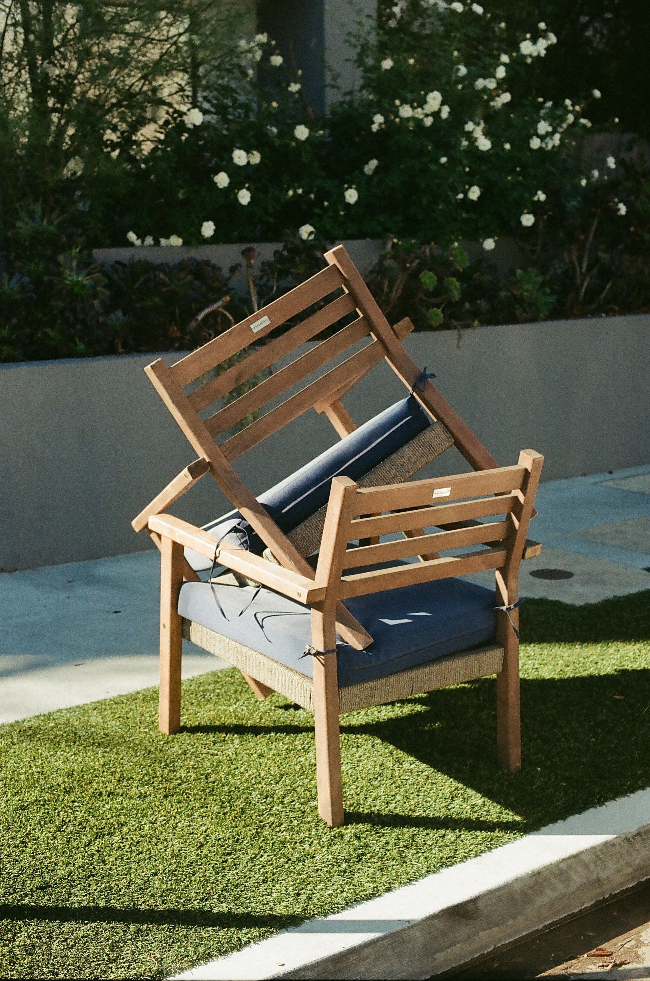 A wooden chair and a corner of a cushioned outdoor sofa on a small patio with artificial grass, with a garden wall and blooming white flowers in the background.