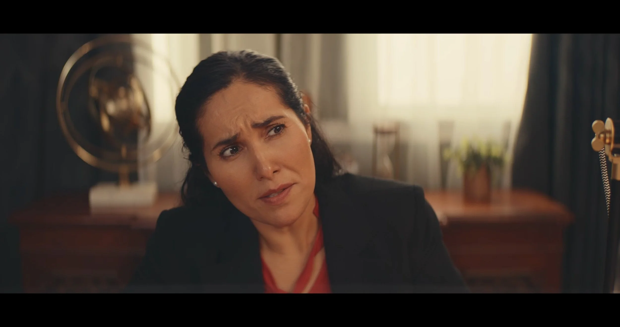A woman with dark hair, wearing a black jacket, looking worried or confused, sitting at a desk with a vintage-style telephone and a fan in the background.
