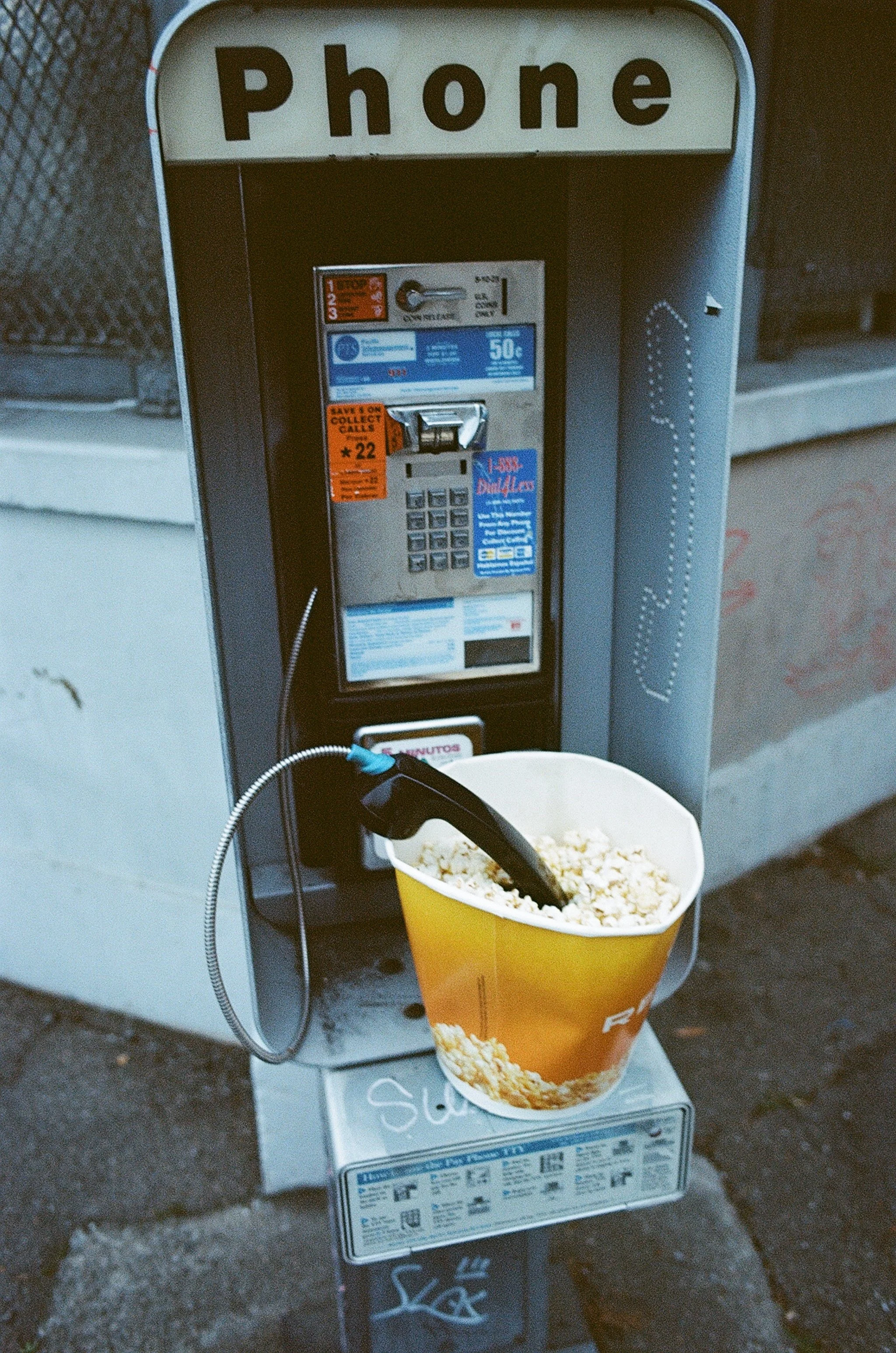 A pay phone on a sidewalk with a bucket of popcorn hanging from the receiver.
