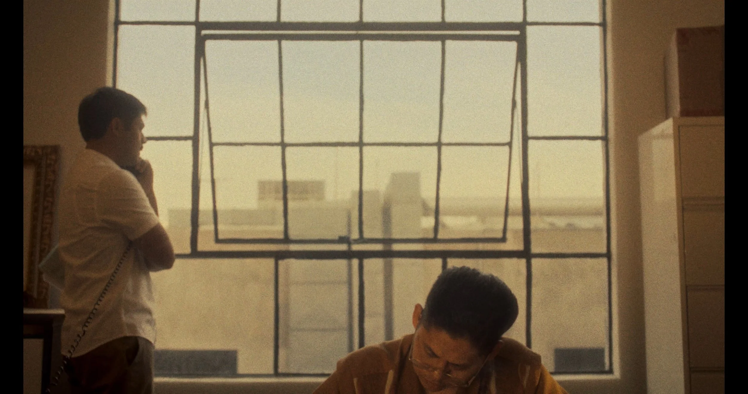 Two men are in a room with large windows in the background. One man, standing on the left, is talking on a telephone and wearing a light-colored shirt. The other man, seated on the right, is looking downward and wearing a brown shirt.