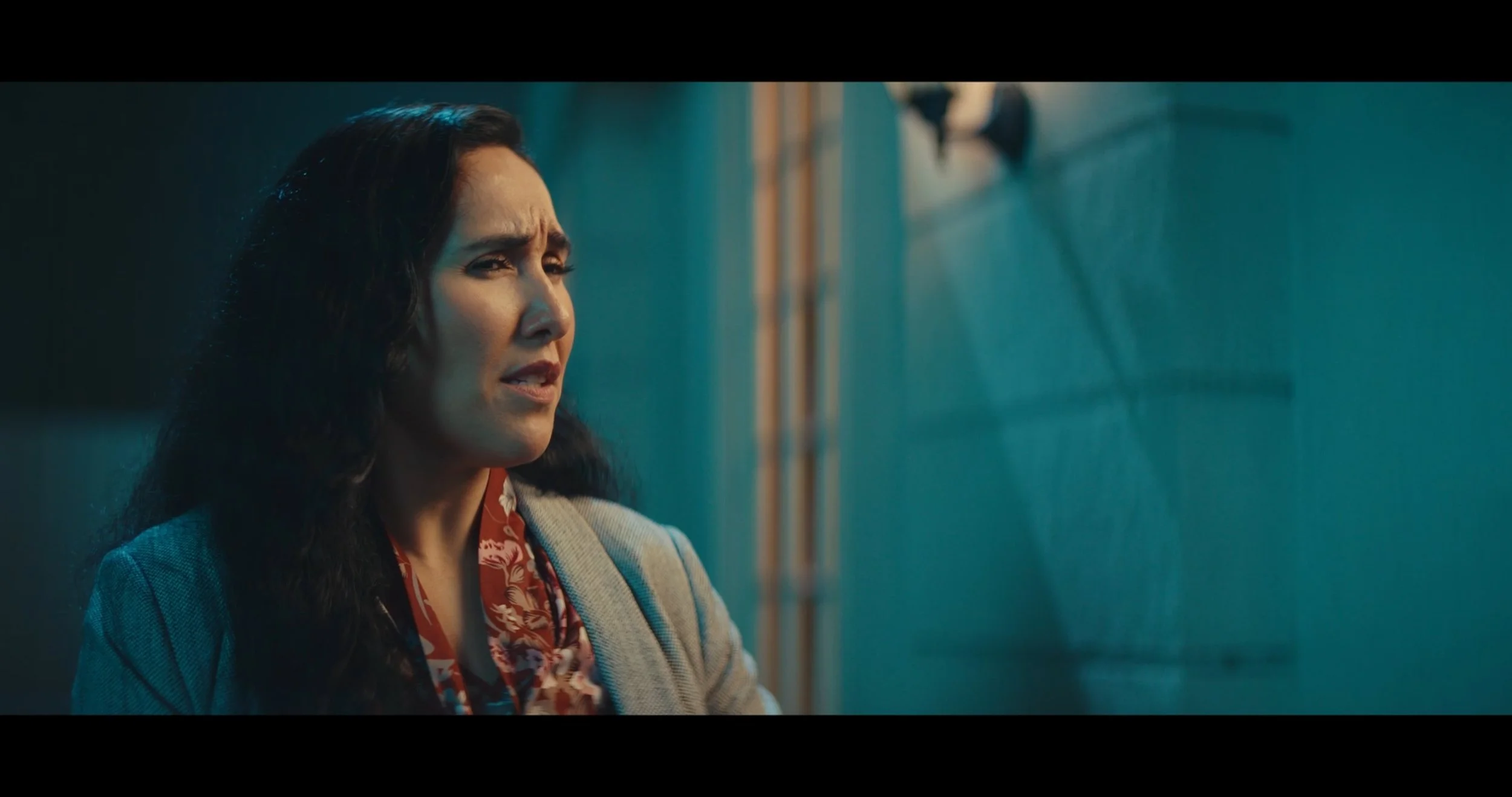 A woman with dark, curly hair, wearing a blue blazer and a red patterned blouse, looks to the side with a concerned or thoughtful expression.