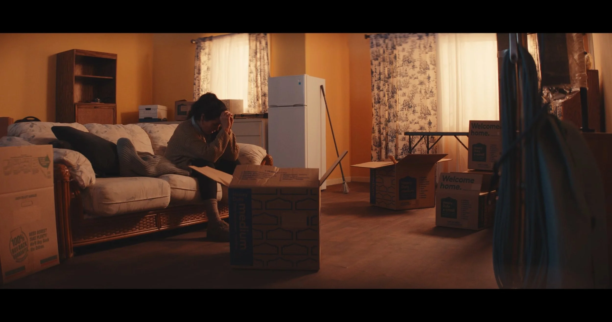 A woman sitting on a sofa in a room filled with moving boxes, looking stressed and holding her head. The room has yellow walls, floral curtains, and sunlight coming through the window.
