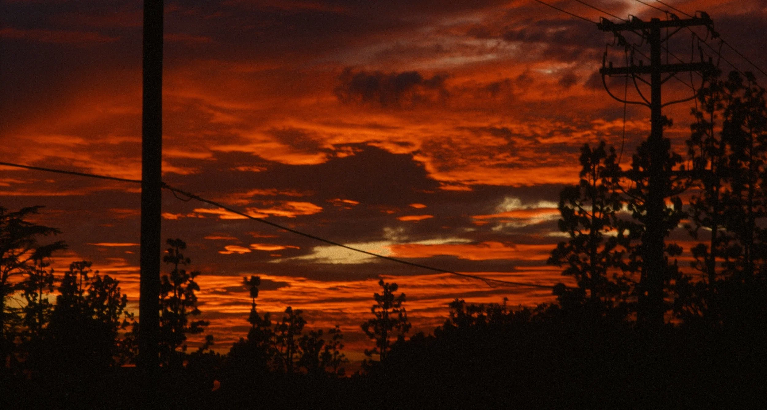 A vibrant sunset sky with orange and dark clouds, silhouetted trees, and utility poles and wires.