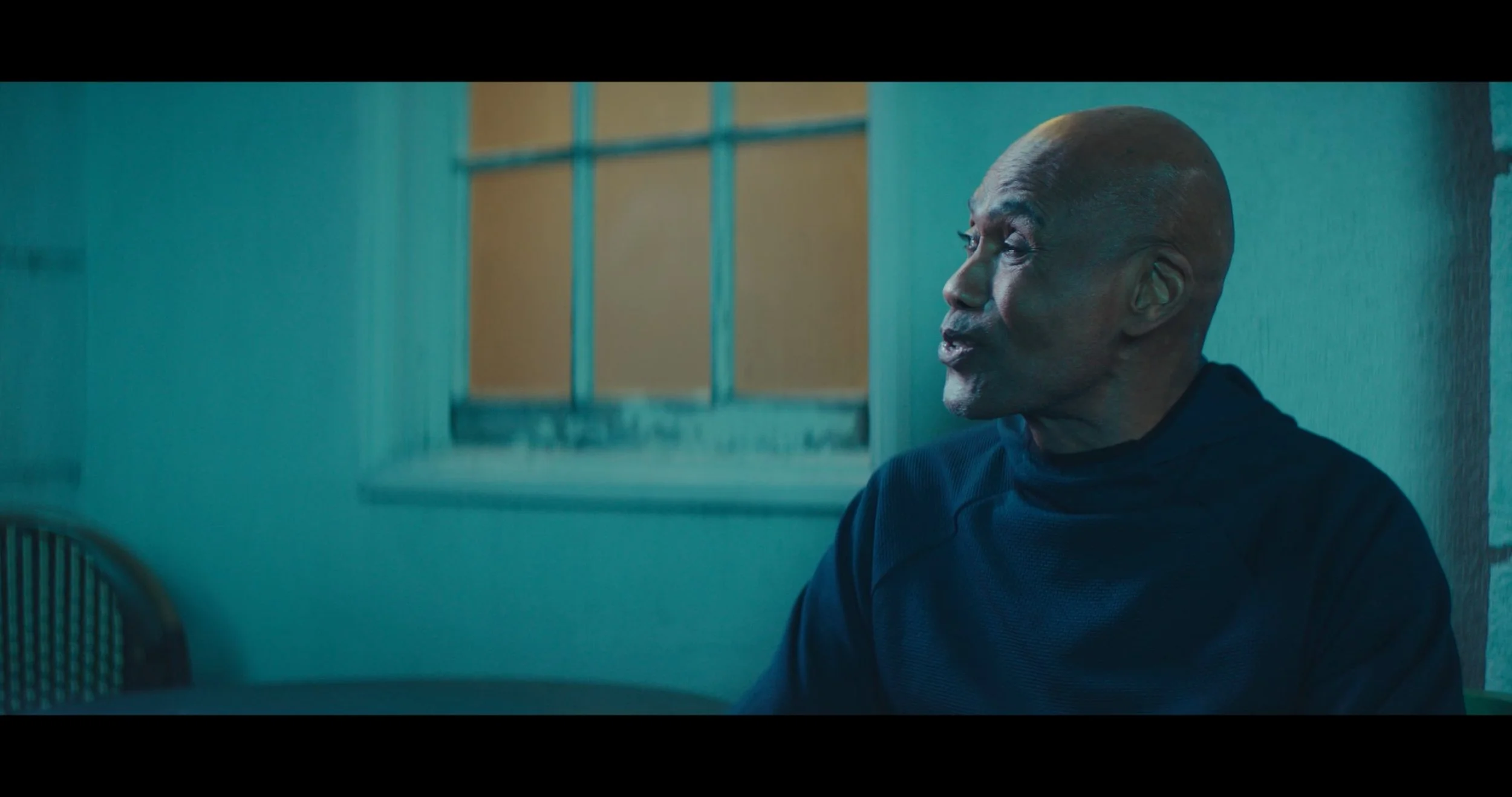 A close-up side profile of an elderly bald man with a dark shirt sitting outside near a weathered wall and window, engaged in conversation.