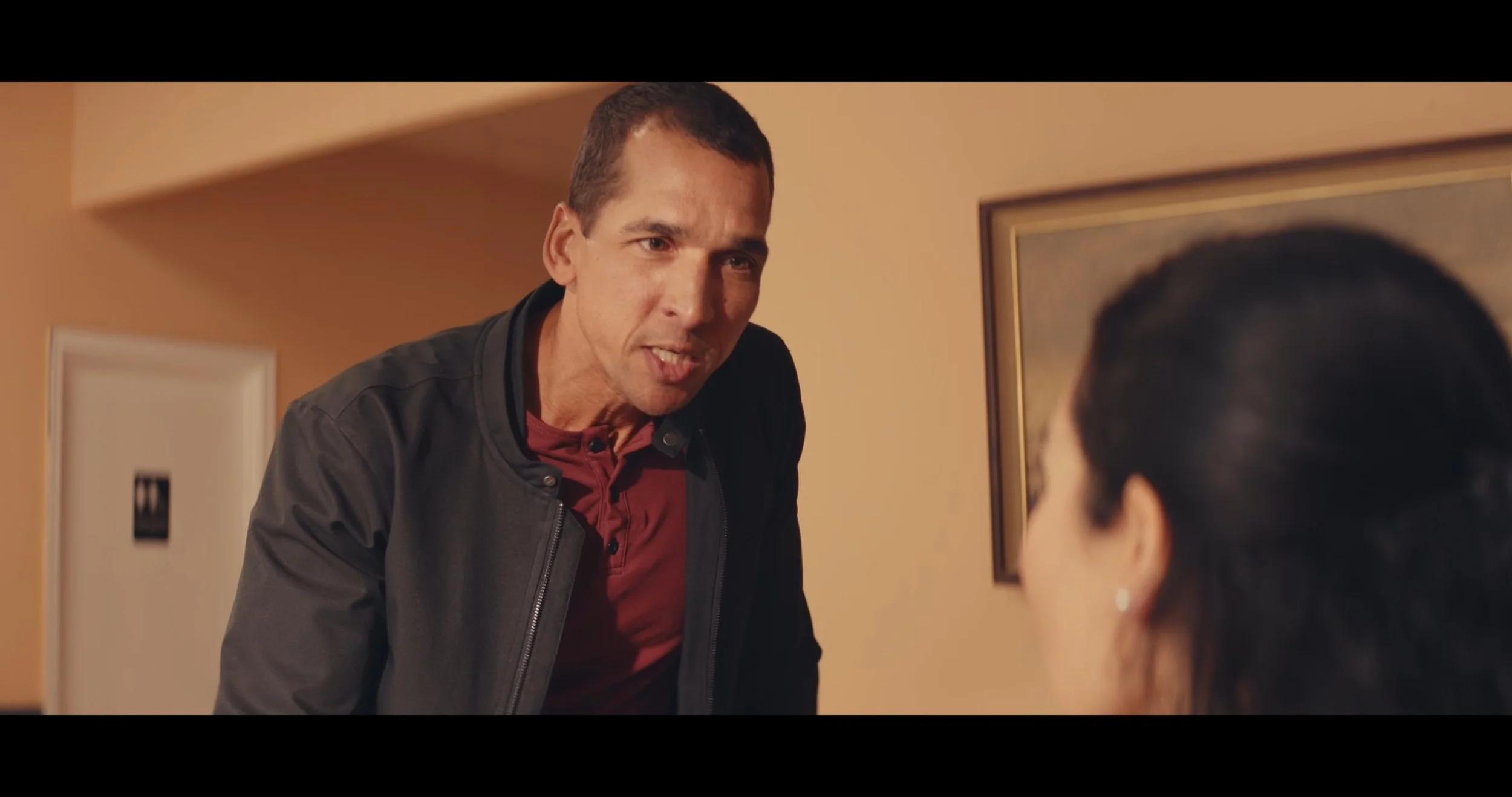 A man is leaning toward a woman who is sitting, in a conversation inside a room with beige walls and framed artwork.