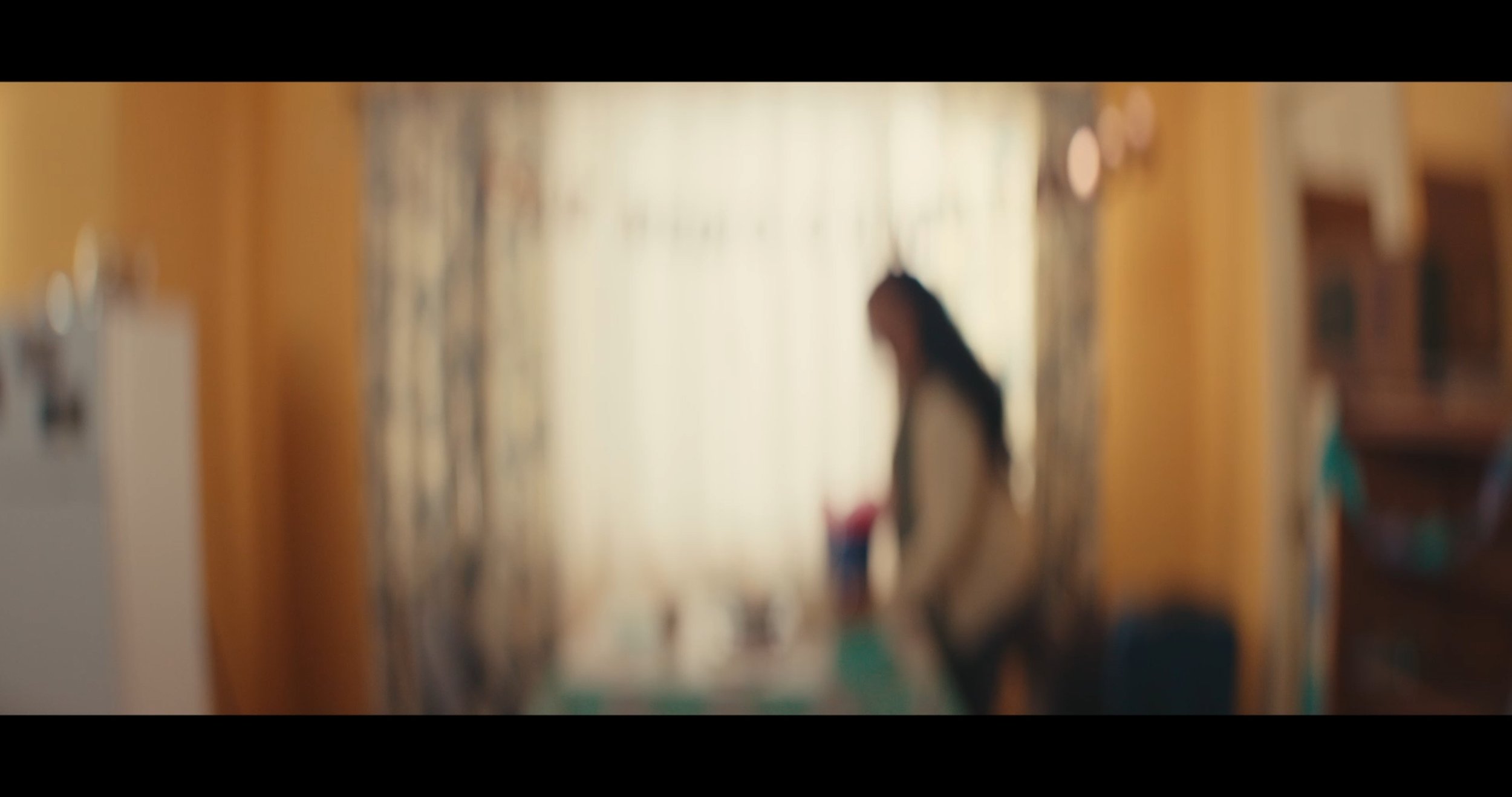 Blurred image of a person standing in a doorway, with a bright background and a yellow wall.