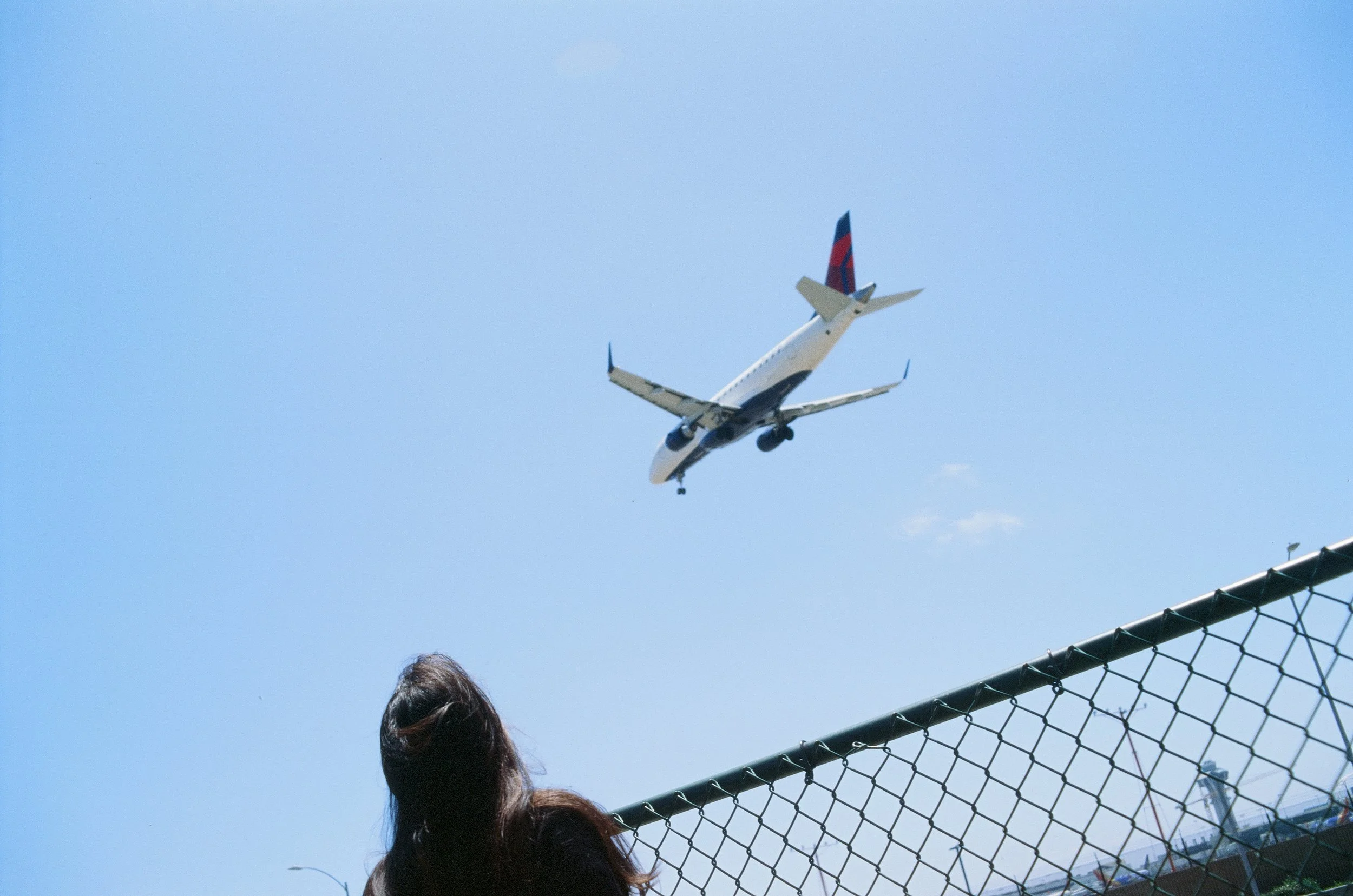 A person with long hair seen from behind watching an airplane flying in the sky above a chain-link fence.