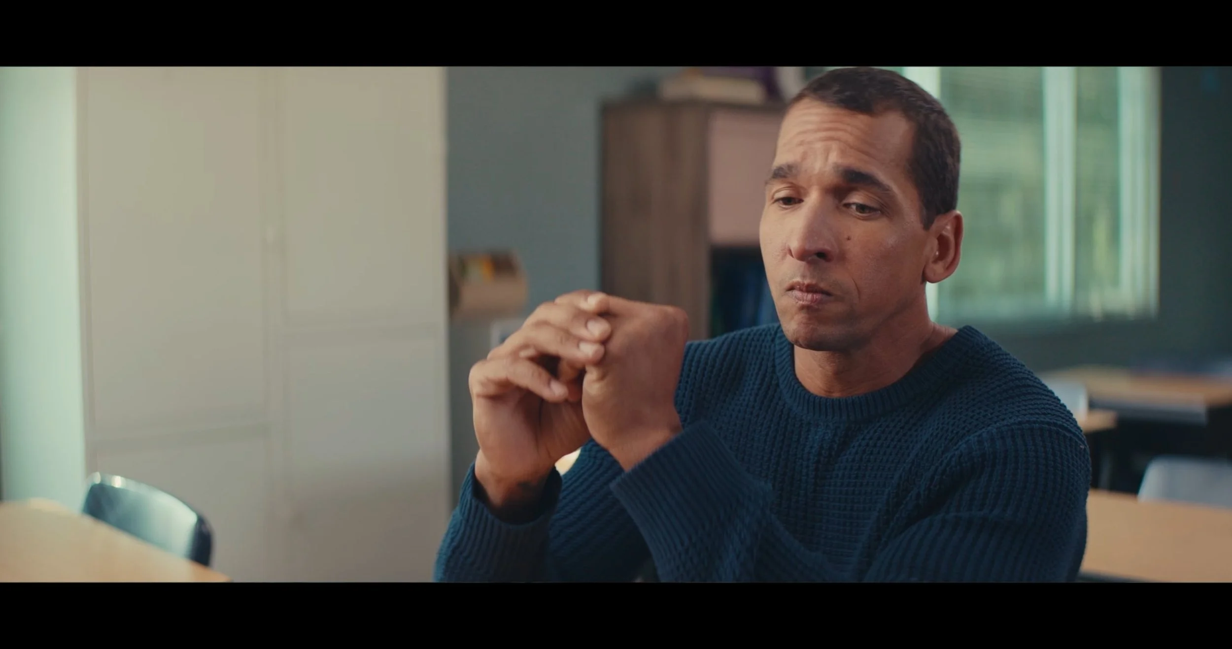 A man with short dark hair and a dark blue sweater, sitting at a desk with his hands clasped and looking thoughtful, in a room with light-colored walls, a bookshelf, and a window with greenery outside.