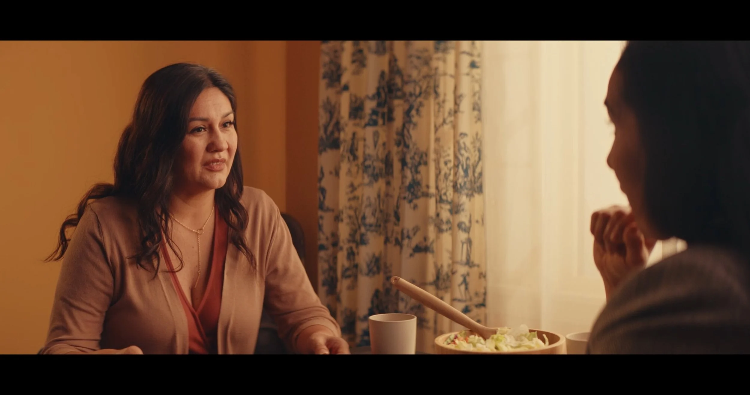 Two women having a conversation at a dining table in a warmly lit room, with curtains and a bowl of salad visible.