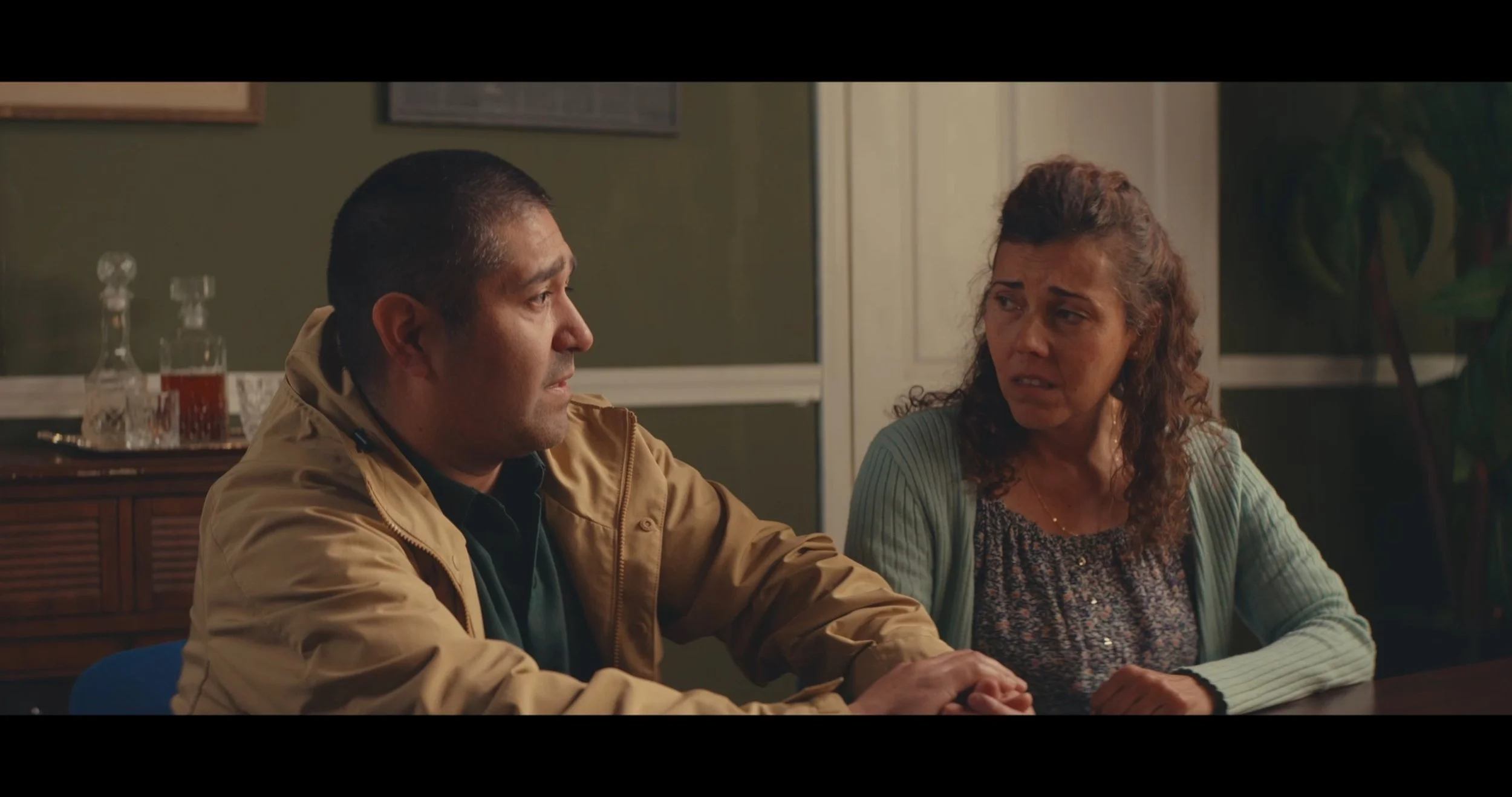 A man and woman sitting at a table having a conversation indoors. The man is wearing a tan jacket and the woman appears emotional.