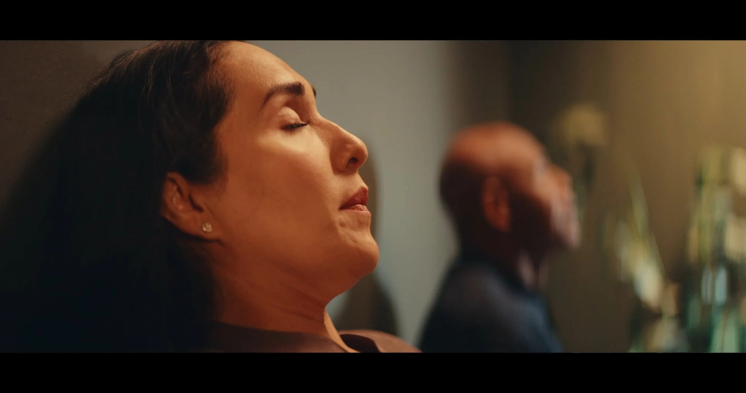 Close-up of a woman with closed eyes, wearing earrings, resting her head against a wall, with a blurred person in the background.