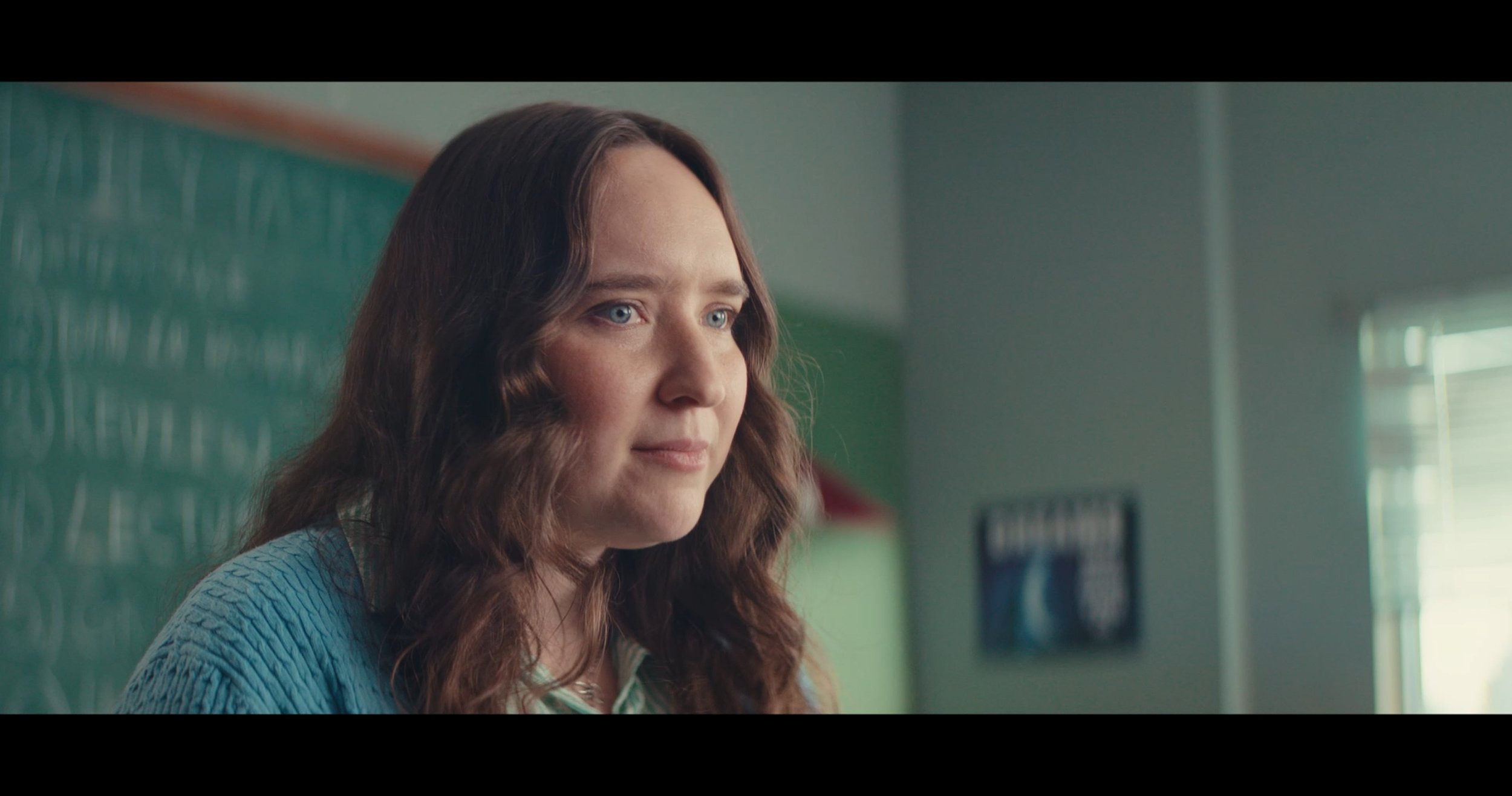 Close-up of a woman with long, wavy brown hair and blue eyes, looking to the right, in a classroom with green walls and windows.