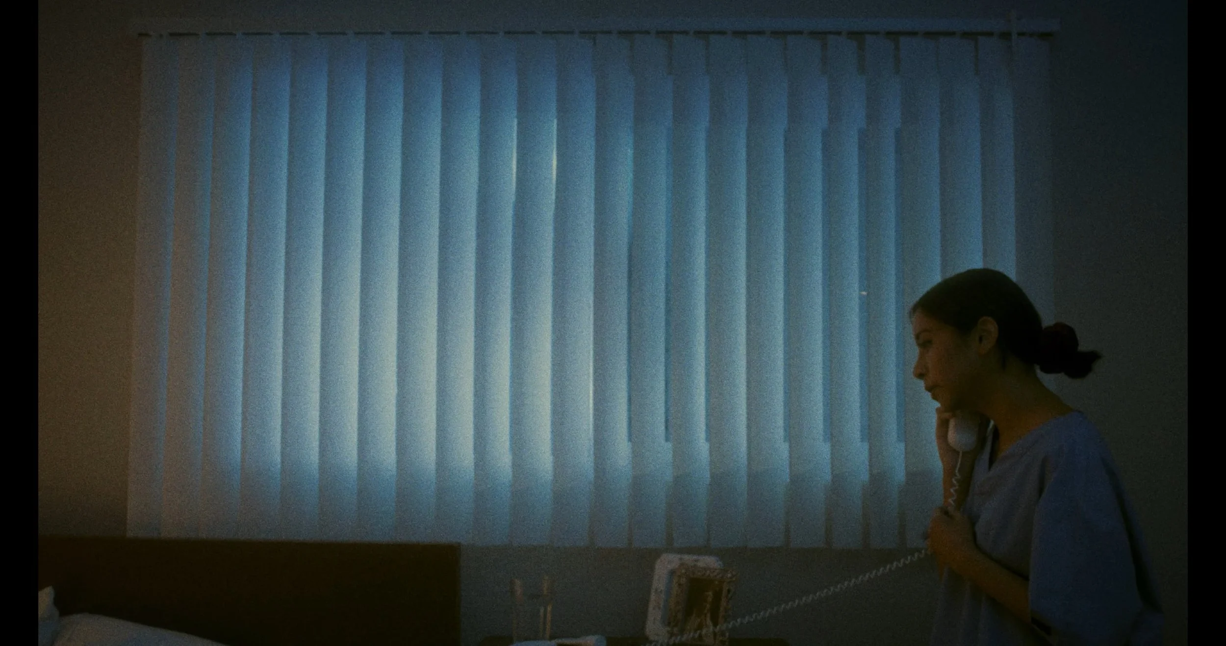 A woman in medical scrubs talking on a landline phone in a dimly lit room with closed vertical blinds covering a window.